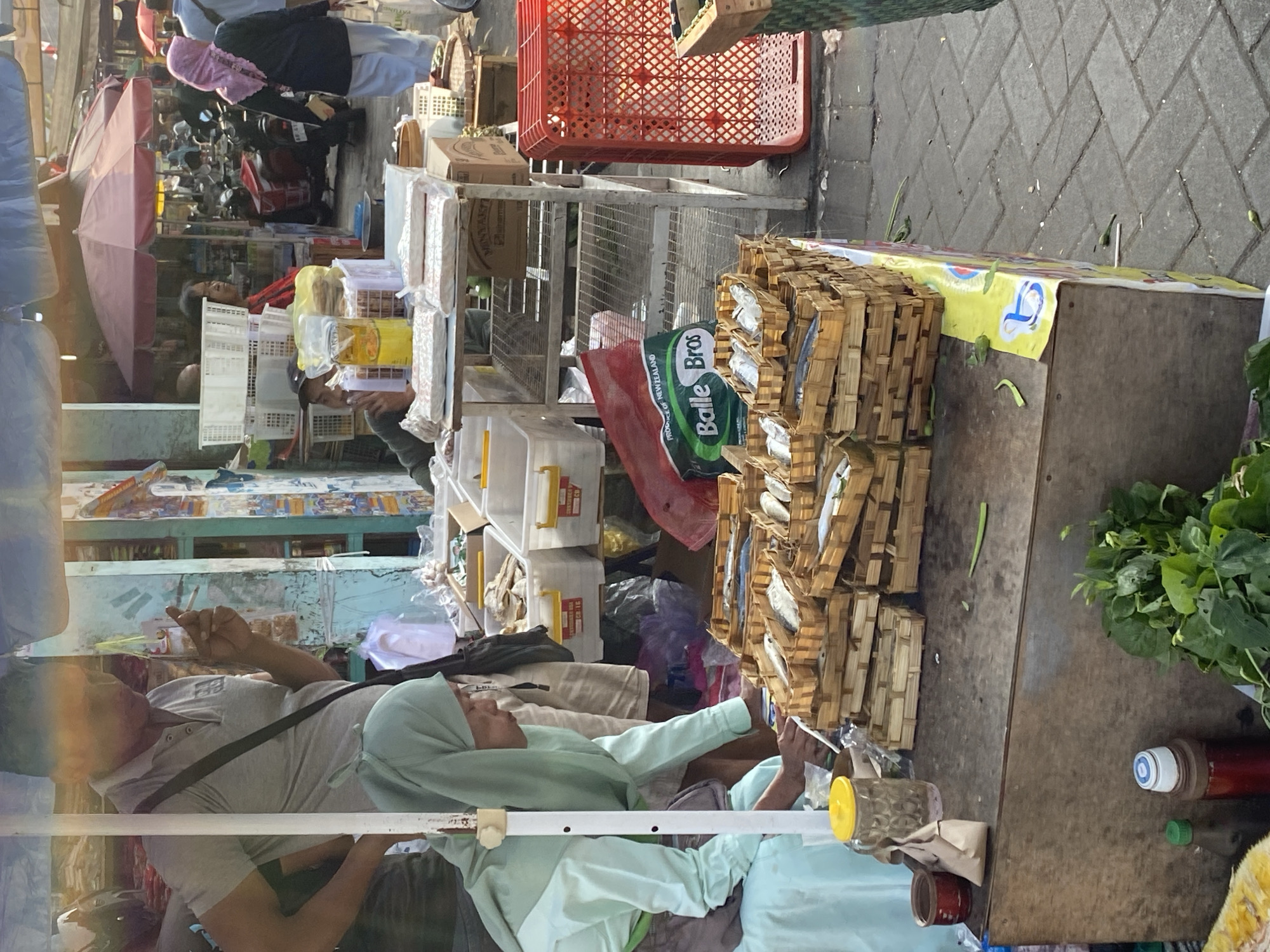 The image captures a bustling outdoor market scene, likely in Ambarketawang, Indonesia, characterized by its vibrant atmosphere and array of goods. In the foreground, a woman wearing a light green hijab and a matching long-sleeved top is bent over a wooden stall, seemingly interacting with the items displayed. The stall is laden with numerous small woven baskets filled with dried fish, arranged in neat stacks. To the left of the woman, a glass jar with a yellow lid and a small paper bag are visible.
Behind her, a man in a grey polo shirt is standing, holding a cigarette in his right hand and appearing to be in conversation. A black strap from a bag is visible across his chest. Further in the background, the market extends with various stalls, people, and vehicles. Red and burgundy umbrellas provide shade over some of the market areas. Motorbikes are parked and some people are seen walking through the market, suggesting active commerce. The ground is paved with grey interlocking bricks. The overall impression is one of a lively and active marketplace where daily trade is taking place.