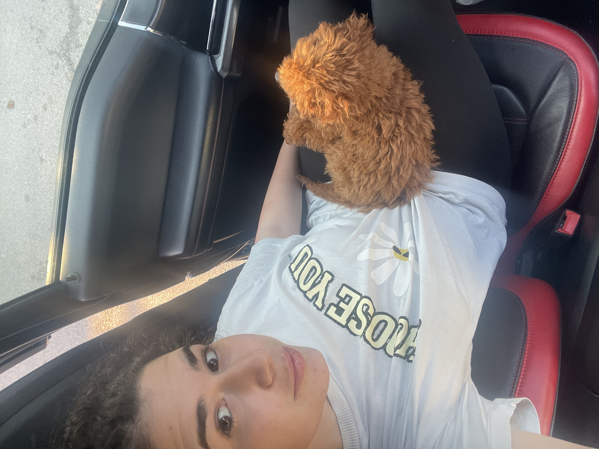 The image, taken in Sofia, Bulgaria, captures a young woman lying back in a car, likely in the passenger seat, with a small, fluffy, reddish-brown dog resting on her chest. The woman has curly dark hair and looks directly at the camera with a calm expression. She is wearing a light-colored t-shirt that features a white daisy graphic and the upside-down text "CHOOSE YOU" in yellow letters.
The small dog, possibly a poodle or doodle puppy, is curled up with its back to the camera, facing away from the woman's head. The car's interior is visible, including a dark seat with red trim and a dark door panel. Bright sunlight streams in through the window, illuminating the light grey pavement outside, suggesting it is daytime, possibly early morning or late afternoon given the warm glow. The scene conveys a relaxed and intimate moment between the woman and her pet inside the vehicle.