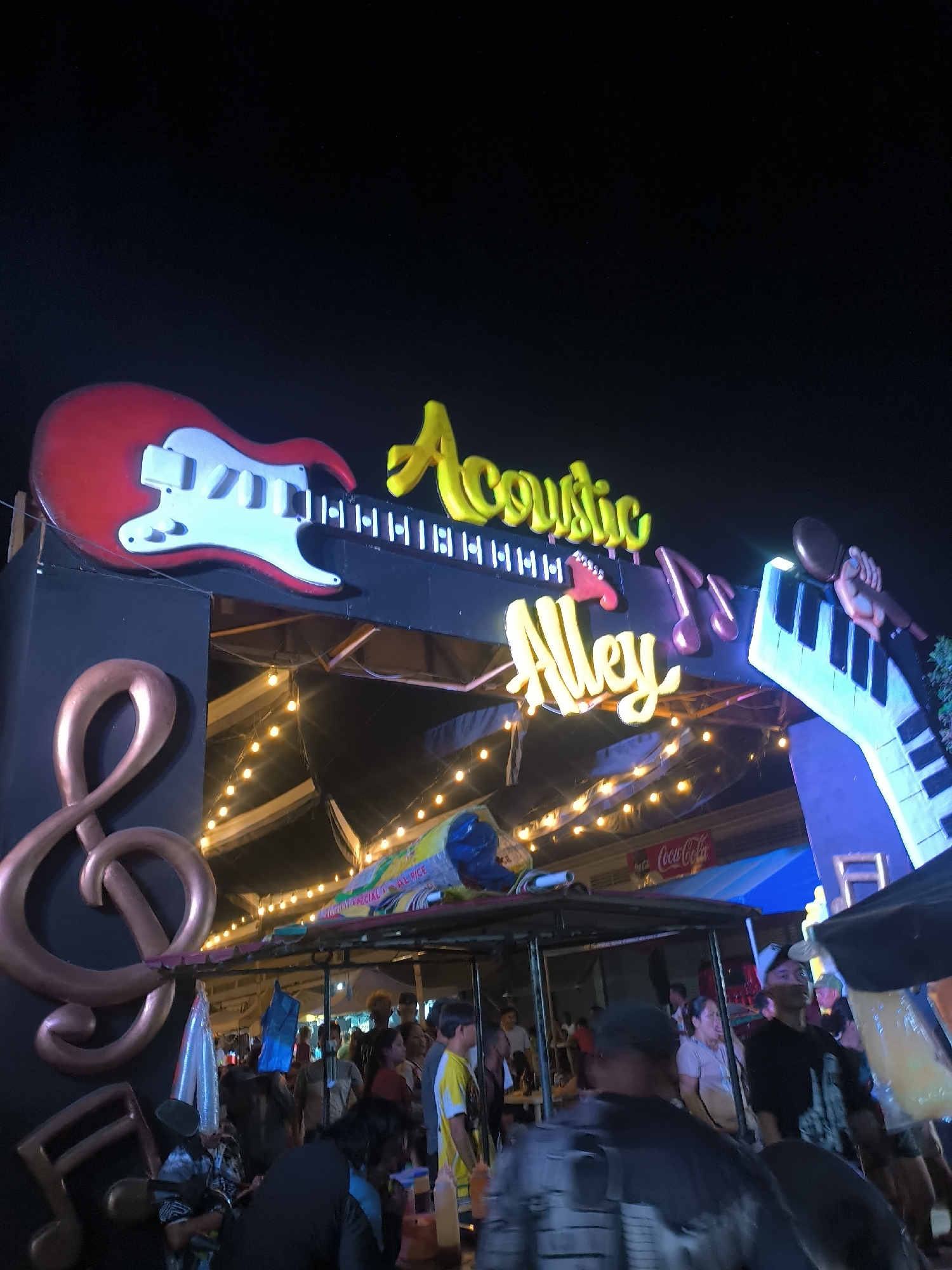 This image captures a lively night scene at "Acoustic Alley" in Kiblawan, Philippines. The foreground is dominated by a large, decorative archway with musical themes. A red electric guitar sculpture is mounted on the left side of the arch, with its strings forming part of the arch's structure. To the right, a keyboard sculpture is depicted with a hand playing it, and musical notes are scattered around the arch. The words "Acoustic Alley" are prominently displayed in bright yellow neon lights across the arch.
Below the arch, a bustling street scene unfolds. Numerous people are visible, some standing and interacting, while others appear to be browsing or walking through. The atmosphere is festive, illuminated by strings of warm, yellow lights that stretch across the area, suggesting an outdoor market or entertainment venue. In the background, tents and stalls are set up, hinting at a lively gathering or festival.
A red Coca-Cola banner is visible in the middle ground, providing a subtle location cue. The time of day is clearly night, indicated by the dark sky and the artificial lighting. The general mood seems to be energetic and cheerful, typical of a night market or a special event. The focus is on the musical theme and the community gathering, creating a welcoming and vibrant ambiance.
