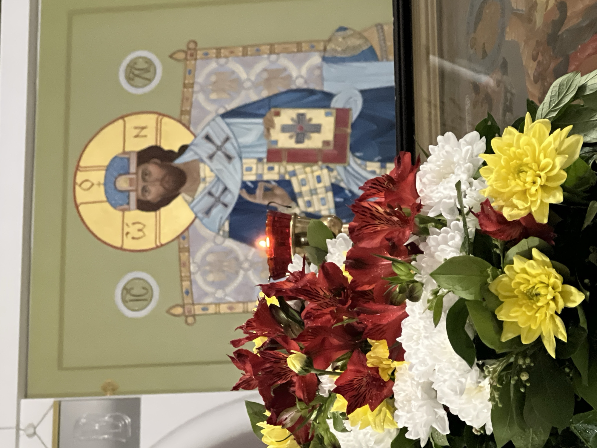 The image shows a religious icon depicting a figure with a halo, dressed in blue and white robes, and holding a book with a cross on it. In front of the icon is a bouquet of flowers in shades of red, white, and yellow. A red candle is visible among the flowers. The icon is framed and mounted on a light green wall with small circular symbols near the top. The location is Khimki, Russia.