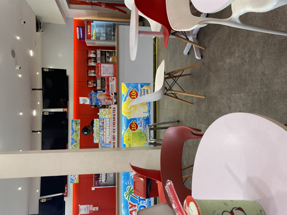 The scene is inside a cafe or restaurant in Kecamatan Piyungan, Indonesia, at the coordinates -7.837 latitude and 110.474 longitude. The cafe has red walls with a white counter separating the customer seating area from the counter. The counter has a sign that reads "INCE 1997 - ICE CREAM & TEA". There are two large, flat-screen televisions mounted on the wall behind the counter. A person, wearing a black cap, stands behind the counter. There are several tables with chairs. One of the tables is positioned closest to the camera, and a half-filled plastic cup containing a green beverage sits on the table. The cup is accompanied by a red plastic straw.
There are four chairs positioned around the table: a red chair with a wooden frame and a white chair with four wooden legs. There are two other chairs: one white and one red, both made of plastic. The chairs are empty. The floor is a mottled grey color.
