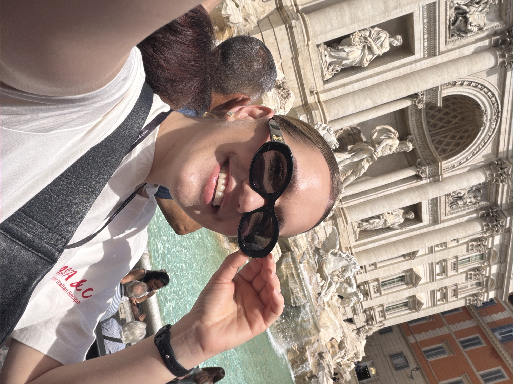 A person is holding sunglasses in front of the Trevi Fountain in Rome, Italy, in a selfie. The person is wearing a white t-shirt with red lettering and a black watch on their wrist. The fountain is a large, ornate structure with sculptures and flowing water. Other people are visible in the background near the edge of the fountain. Buildings are seen behind the fountain.
