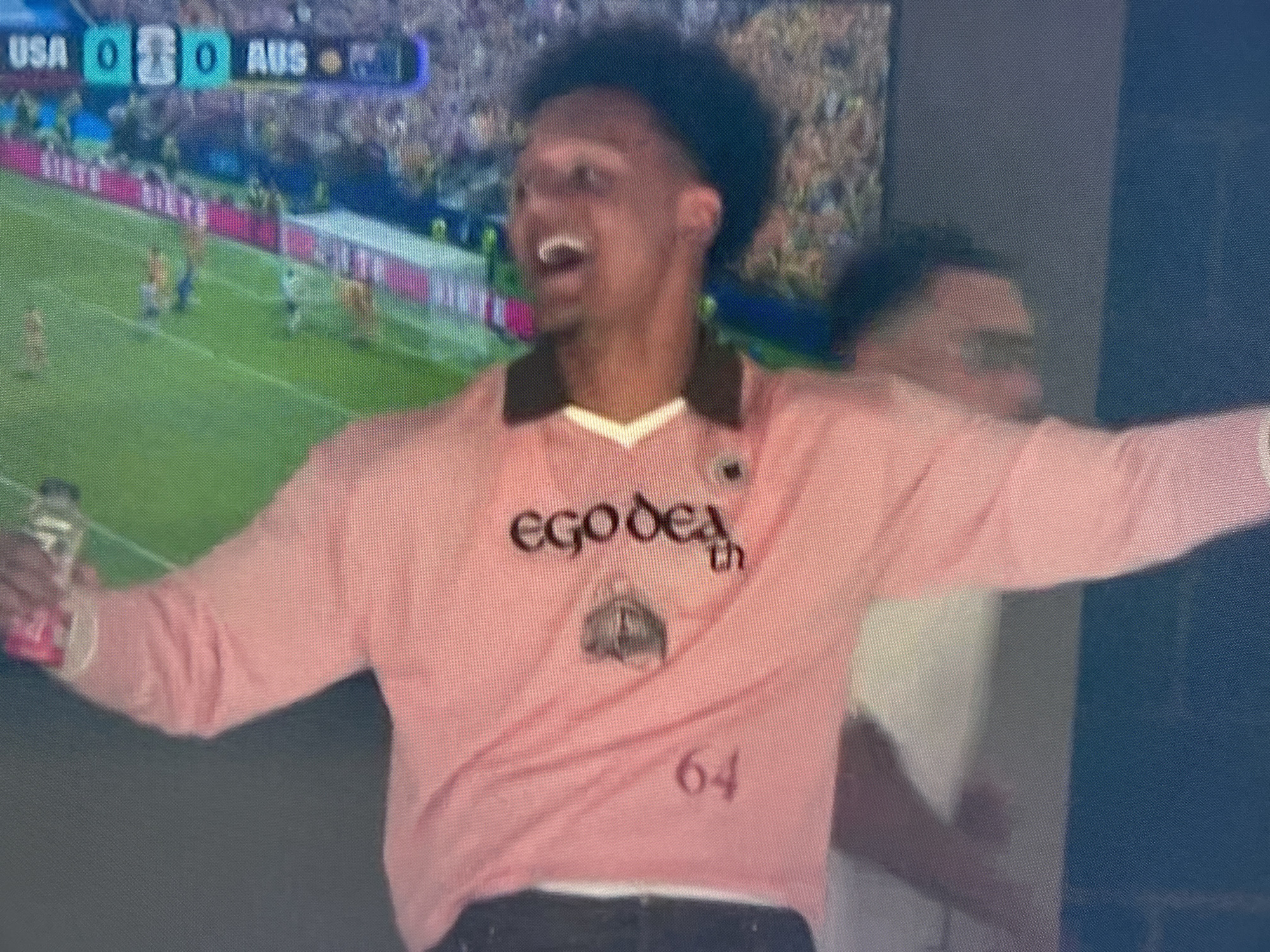 A man wearing a pink long-sleeved shirt with the words "ego death" and the number "64" printed on it is seen in the foreground. He is holding a bottle in his left hand and appears to be cheering or reacting to something off-camera. In the background, a scoreboard displays "USA 0 0 AUS", indicating a soccer match is being watched. The overall setting is likely an indoor venue with a large screen showing the game, possibly a bar or a private viewing party.