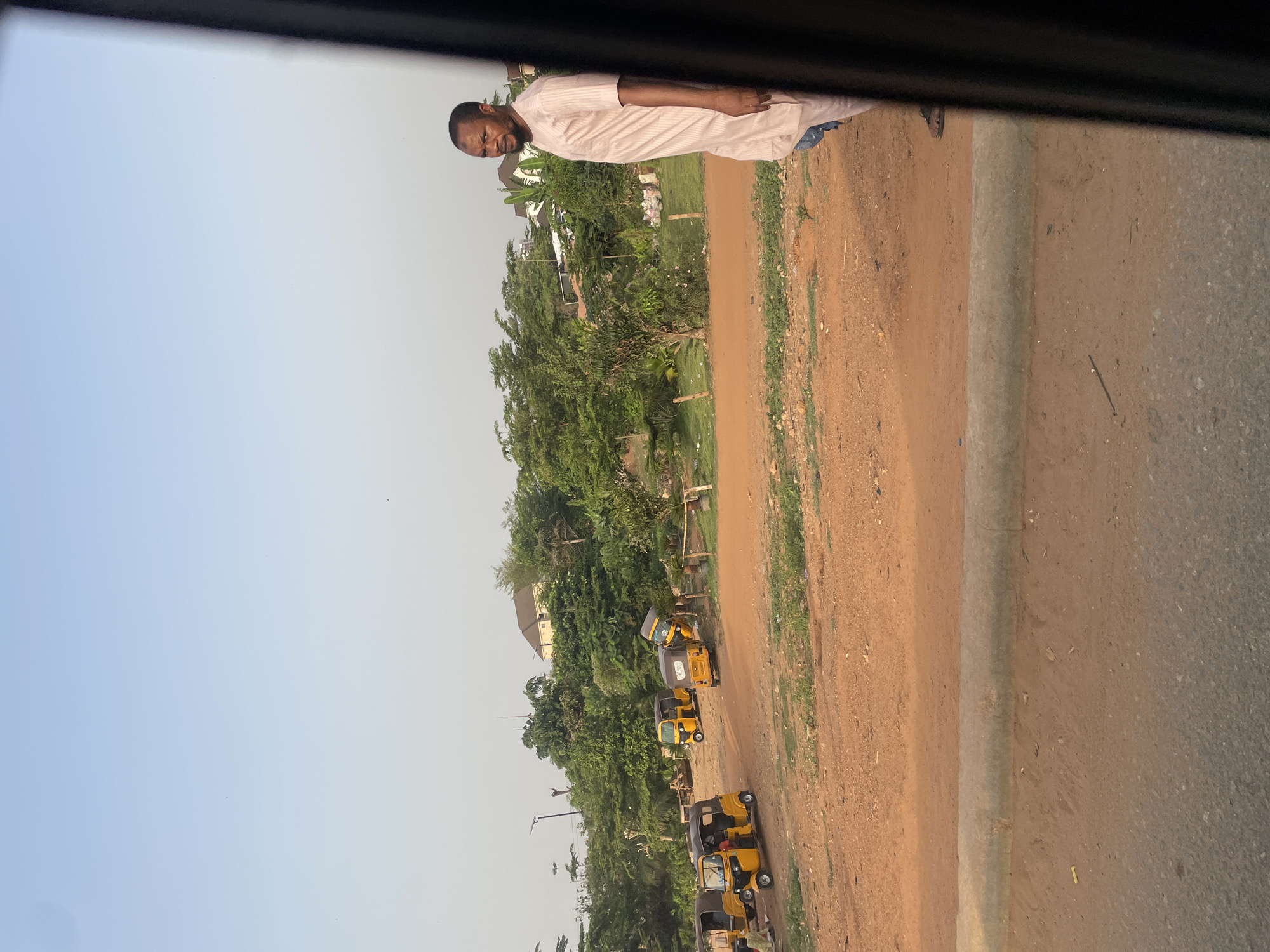 The image, rotated 90 degrees clockwise from its original orientation, depicts an outdoor scene in what appears to be a semi-urban or suburban area, consistent with Abuja, Nigeria.
The primary subject is a dark-skinned man standing on a reddish-brown dirt path to the left of the frame. He wears a light-colored, possibly striped, long-sleeved tunic, resembling a traditional Nigerian caftan or agbada top, over light blue trousers. He has a beard and looks directly towards the viewer with a neutral expression.
Behind the man and extending further into the background is a row of several yellow and gray auto-rickshaws, commonly known as Keke Napep or Maruwa, parked along the dirt path amidst dense green trees and vegetation. Scattered among the greenery, residential buildings are partially visible in the distance.
In the immediate foreground, a concrete curb separates the dirt path from a paved road, suggesting the image was taken from a vehicle looking out a window, indicated by the dark frame visible at the top and bottom edges (when re-oriented). The sky is clear and bright, ranging from light blue to a pale white, indicating either early morning or late afternoon. The overall weather appears sunny and calm. No visible text is present in the image.