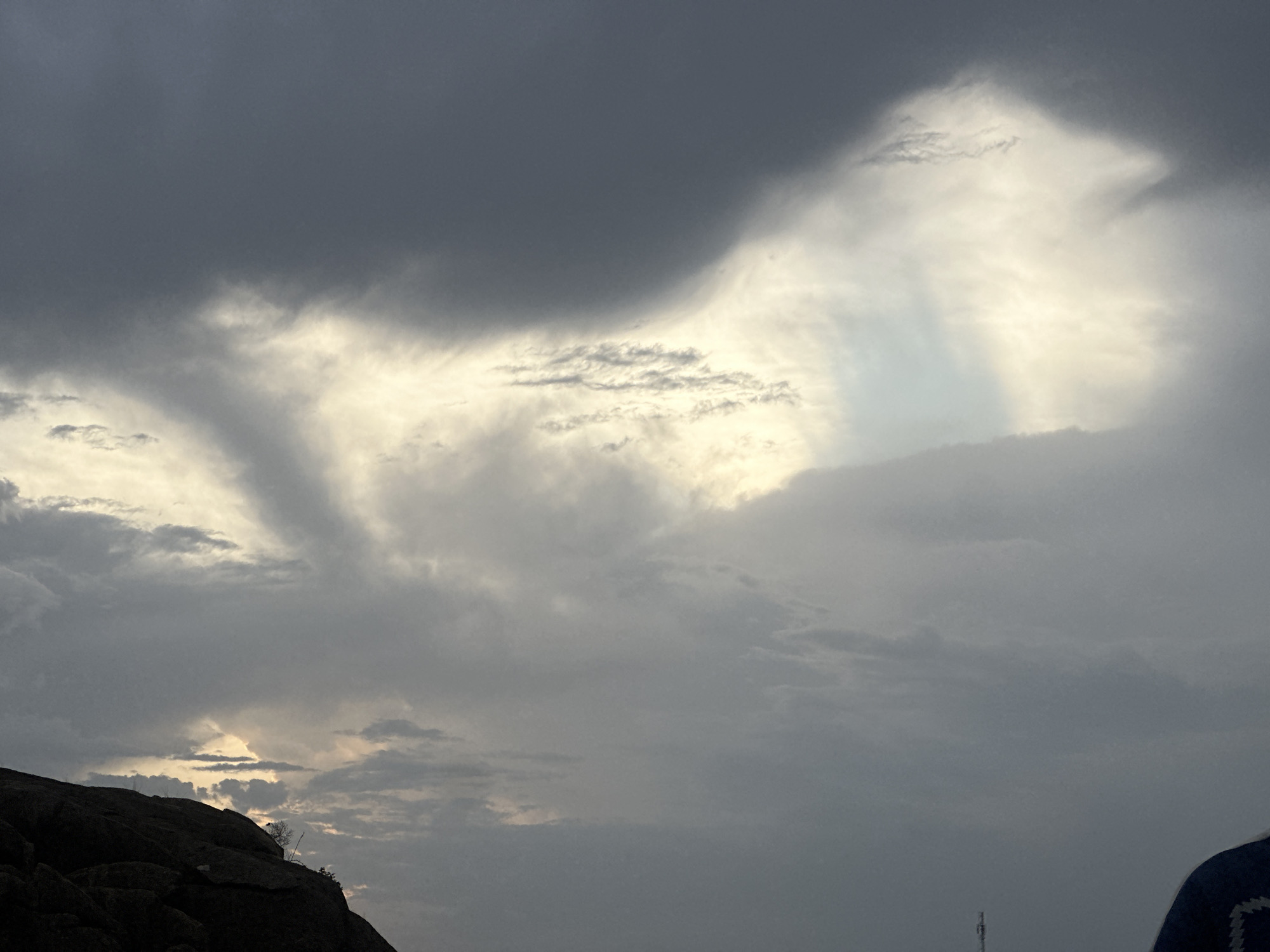 The image shows a cloudy sky above a rocky outcrop. The sky is predominantly gray with patches of lighter, white clouds. Sunlight filters through some of the clouds. In the lower left corner, a dark, rocky outcrop with some vegetation is visible. A tower is slightly visible on the bottom right. The location is Jos, Nigeria.
