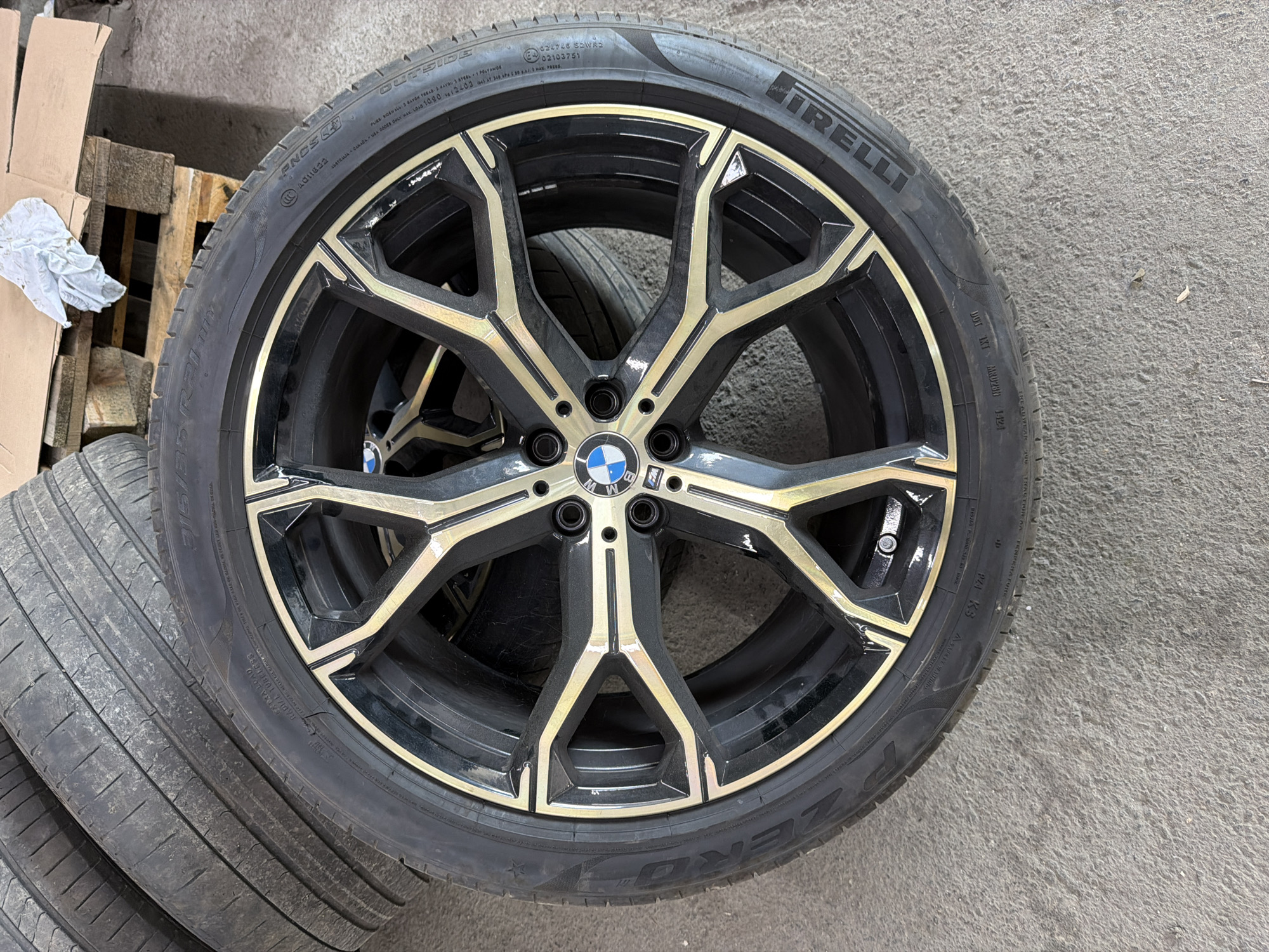 The image captures a single car wheel and tire, likely in a storage or workshop environment in Burgas, Bulgaria. The wheel is a multi-spoke design, characterized by a black finish with striking gold or brushed metal accents on its spokes. A prominent BMW logo is centrally placed on the wheel.
Mounted on the wheel is a Pirelli P Zero tire, clearly identified by the "PIRELLI" and "P ZERO" branding on the sidewall, accompanied by a star symbol indicating its approval as original equipment for BMW vehicles. Key details visible on the tire include its size "245/35 ZR21", along with designations such as "OUTSIDE", "PNCS", "EXTRA LOAD", and "RADIAL TUBELESS". Manufacturing origin "MADE IN ROMANIA" is also present. Further technical specifications detail the "MAX. LOAD 1090 KG (2403 LBS) AT 300 KPA (44 PSI) MAX. PRESS. COLD" and ply composition. Various certification marks like "E4 02103751" and "DOT MKT MR028N 1421", as well as "PZ4" and "K3", are visible.
In the background to the left, additional used tires are stacked, partially out of view. Wooden pallets and cardboard boxes further suggest a storage or industrial setting. The ground is a uniform concrete surface. The lighting is even, implying daytime conditions, possibly indoors or under an overcast sky. There is no activity taking place, and no specific time of day or emotional cues are evident.