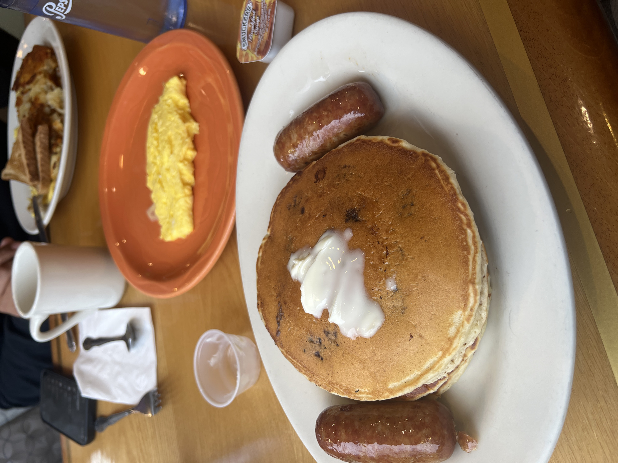 The image captures a breakfast scene, likely in Balmville, United States of America, given the origin of the image. The focus is on a white plate containing a stack of pancakes topped with a dollop of white butter and accompanied by two sausages. To the left, there is an orange plate holding scrambled eggs, a white plate containing hash browns and toast, and a white mug near cutlery on a napkin. A bottle of Pepsi and a small container of Smucker's syrup are also present. The scene is set on a wooden table. There is a person visible to the left, partially captured in the frame. The lighting suggests an indoor setting.