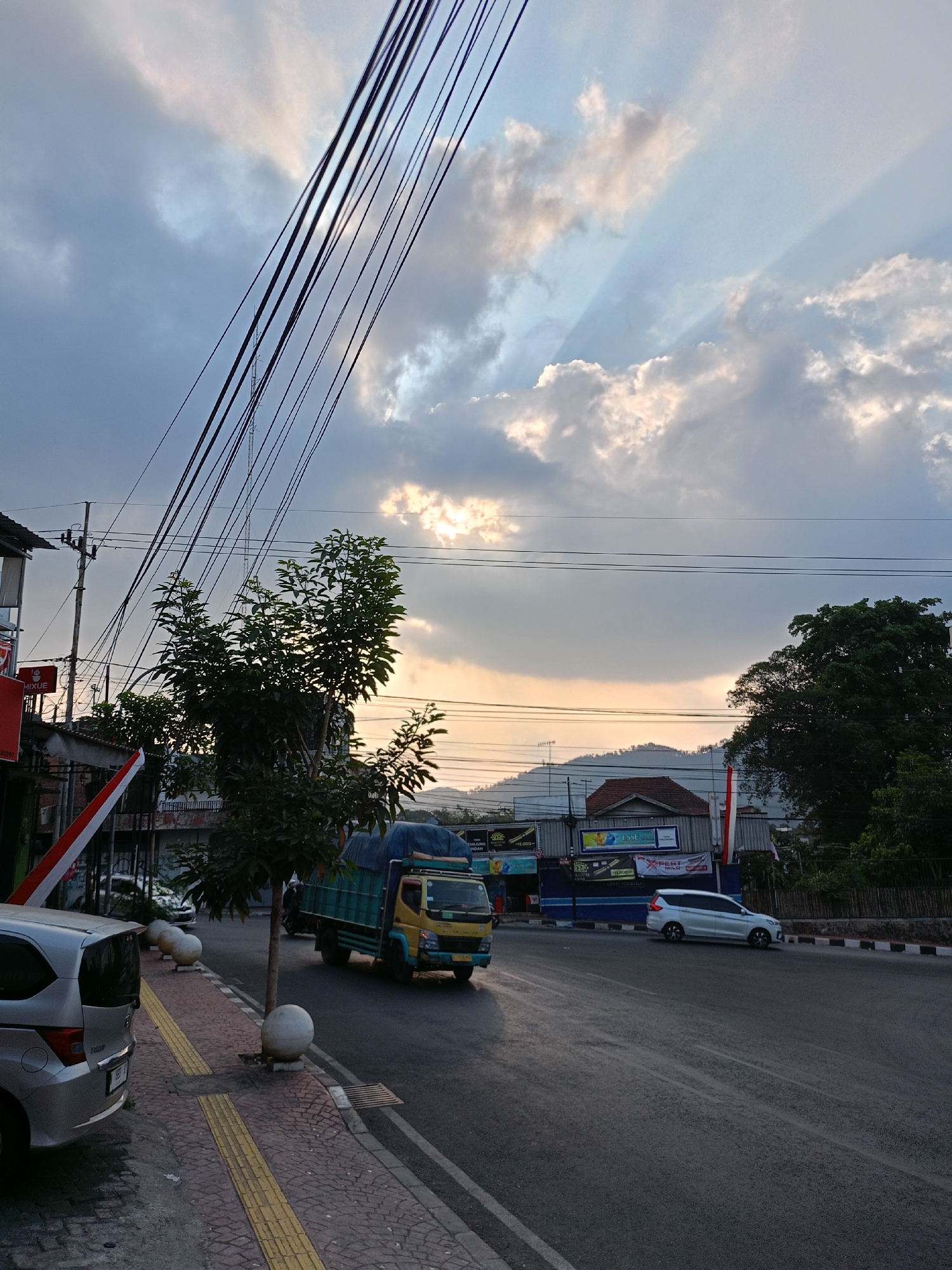 The image captures a street scene in Batu, Indonesia, during what appears to be late afternoon or early evening. The sky is dramatic, with dark clouds partially obscuring the setting sun, casting rays of light through the gaps. Overhead, a dense network of power lines crisscrosses the frame, creating a stark visual element against the sky.
In the foreground, a silver hatchback is parked on the left side of the street, its rear visible. Adjacent to it is a sidewalk with yellow tactile paving. A young tree stands on the sidewalk, its branches reaching upwards. Further down the sidewalk, large spherical bollards are positioned.
The street itself is paved and shows signs of traffic. A yellow and green truck with a covered load is driving on the road, heading away from the viewer. A white MPV is also seen driving on the road, moving in the same direction as the truck.
On the right side of the road, there are buildings, some of which appear to be shops or businesses with signage. One building has a prominent red and white flag displayed vertically. Behind the buildings, a tree-covered hillside is visible, with a roof of a house peeking through. The overall impression is a busy urban street in a tropical environment, with the natural beauty of the sky and landscape contrasting with the man-made infrastructure.