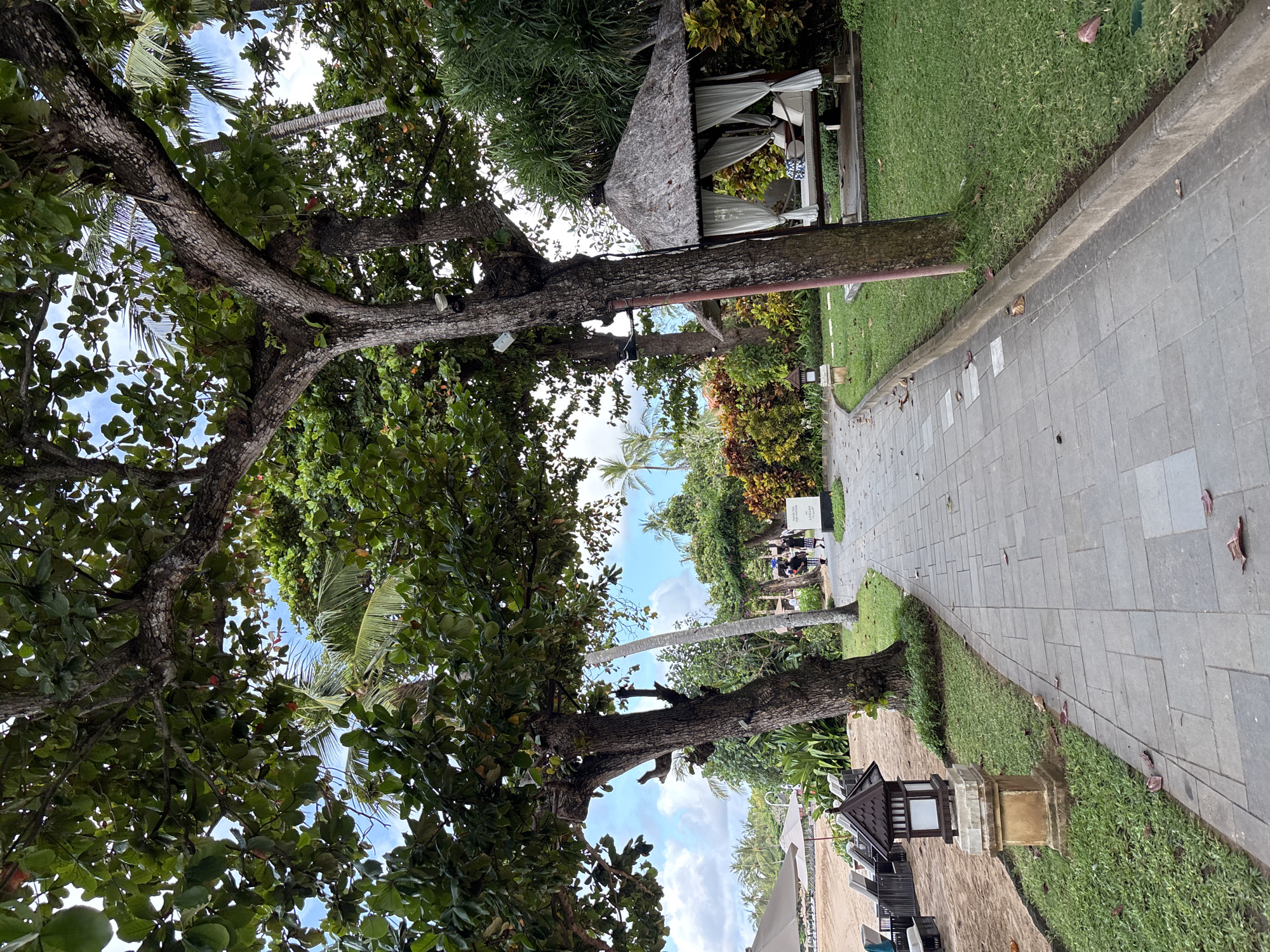 The image captures a view in Kuta Selatan, Indonesia, with a blend of natural and constructed elements. A paved walkway stretches into the distance, bordered by manicured grass. On the left, mature trees dominate the scene, their canopies shading the area. Further along the path, two people walk away from the camera. A thatched-roof structure is situated on the grass. In the foreground, near the path, is a small, decorative lantern-like structure. In the background, the beach is visible.