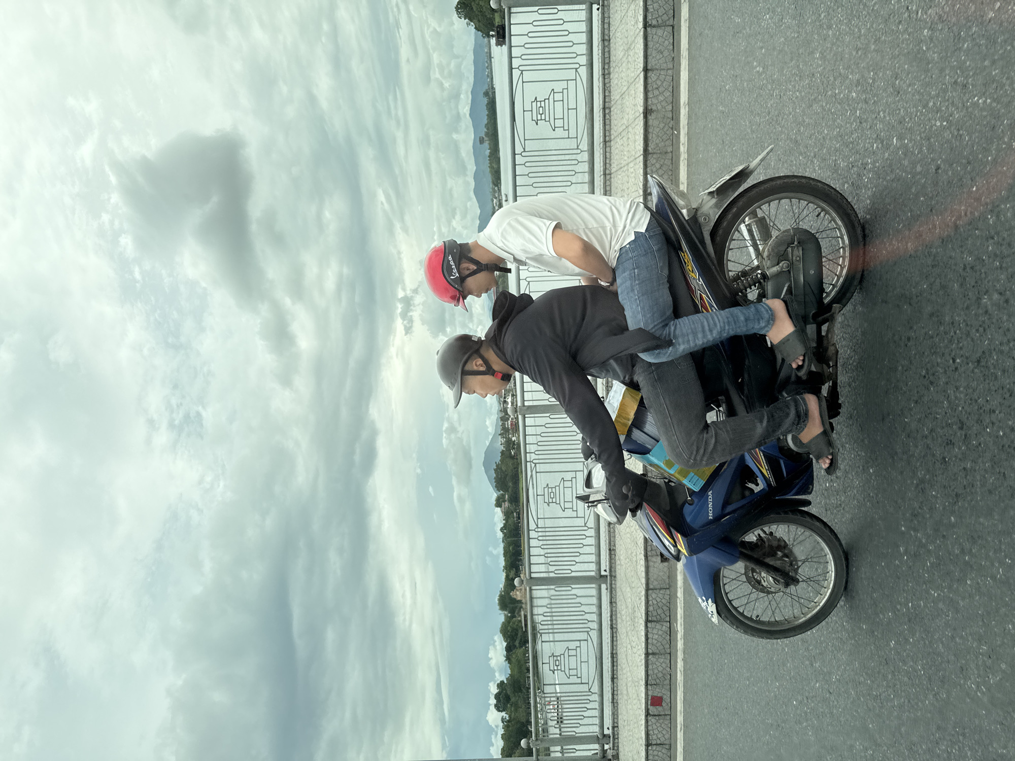 Two individuals, both wearing helmets, ride on a blue Honda motorcycle on a paved road, possibly a bridge given the railing and water visible in the background. The rider in front wears a dark jacket and a grey helmet, while the passenger behind wears a white polo shirt, blue jeans, and a red helmet. They are moving from left to right in the frame, with the motorcycle angled slightly towards the viewer. The background features a body of water, a decorative metal railing with pagoda-like motifs, and distant trees and hills under a cloudy sky. The scene appears to be in Thuận Hóa Ward, Vietnam, based on the presence of a Honda motorcycle and the characteristic architecture of the railing.