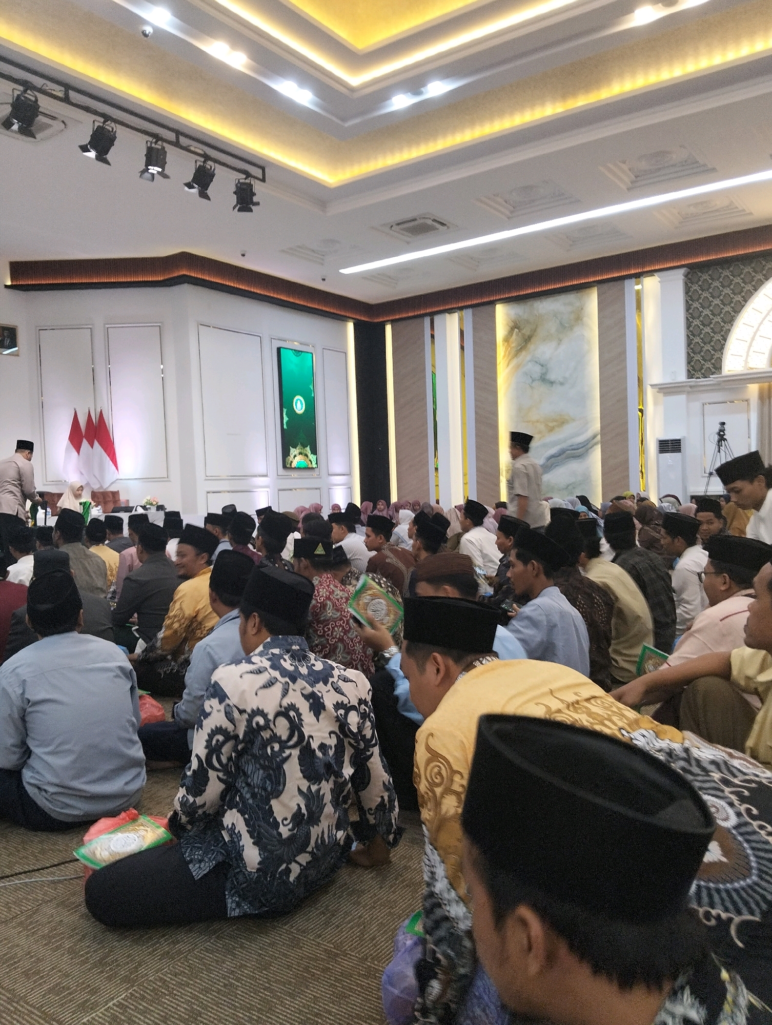 The image depicts an indoor gathering of people, likely a religious event given the attire and the presence of flags resembling the Indonesian national flag in the background. The room is well-lit with recessed ceiling lights and spotlights, and features decorative elements on the walls, including marble-like patterns and patterned panels. Many individuals are seated on the floor, some wearing traditional Islamic caps (peci). The central focus is on the gathering itself, with people looking towards a raised area or stage that is partially visible. The overall atmosphere appears to be solemn and respectful. The image was taken from a low angle, looking upwards towards the ceiling and the gathered crowd. The location is identified as Dagan, Indonesia.