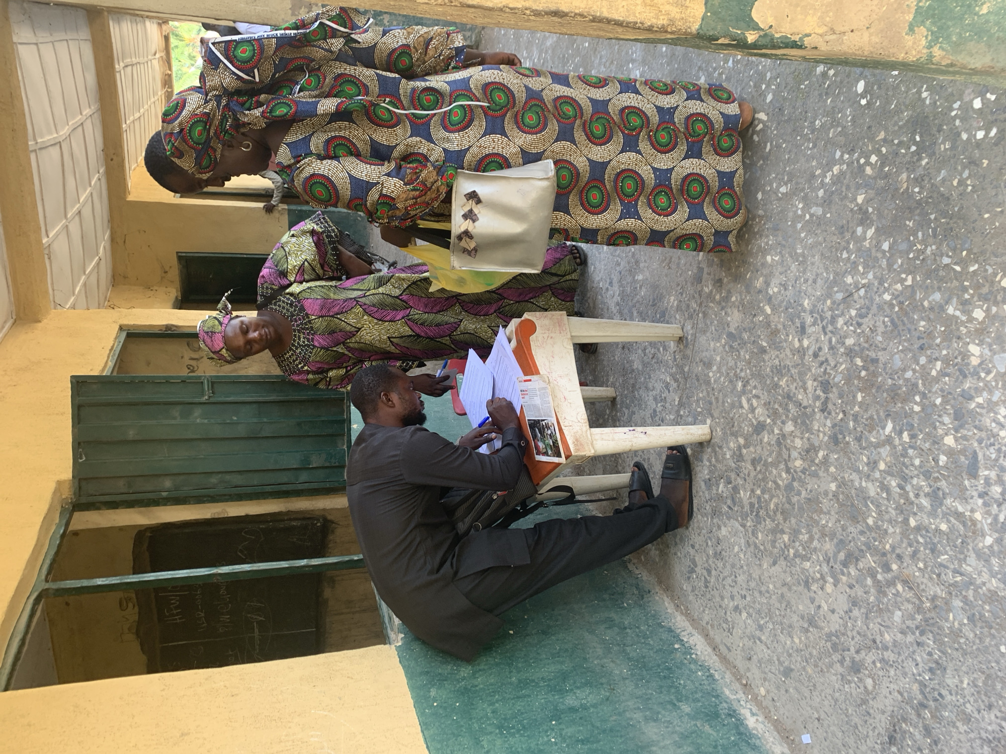 The image shows three people in what appears to be a polling station in Andaha, Nigeria. Two women wearing brightly patterned dresses and head wraps stand near a man who is seated at a small table. The man is dressed in a dark suit and is writing on a piece of paper. Papers and a clipboard are on the table. A handbag rests near the table. The location seems to be outdoors but adjacent to a building with yellow walls and green window frames. The ground is a patterned concrete. Writing is visible on a dark chalkboard-like surface visible through a window.