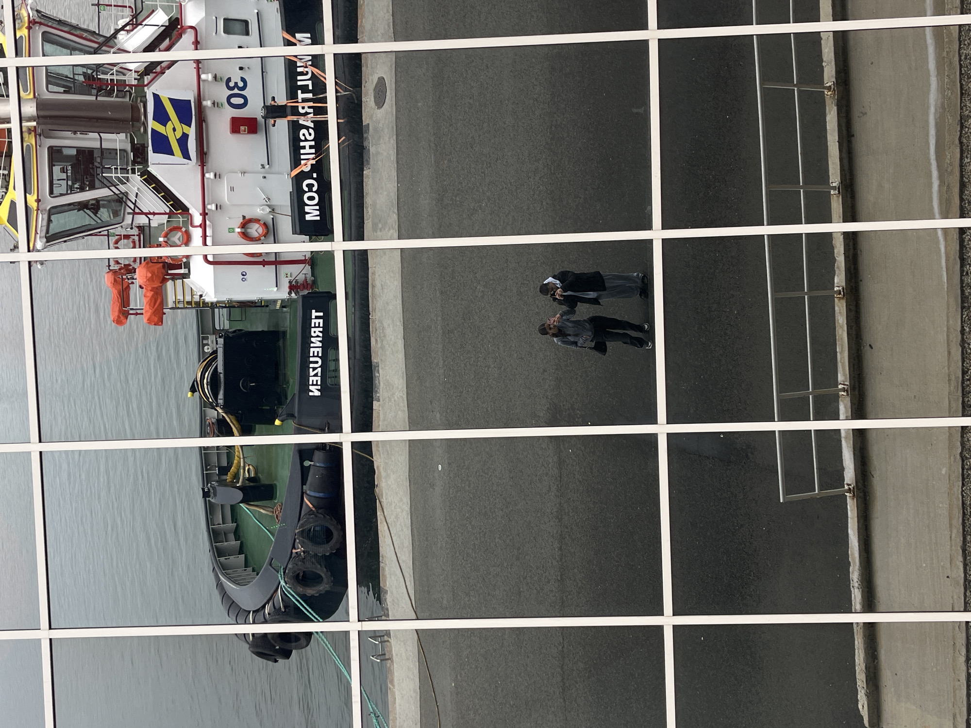 The image shows a reflection in a building's window. The reflection includes a section of the exterior: a tugboat docked in the water next to a concrete pier, and two people standing on the pier, one pointing a camera toward the water. The side of the tugboat shows the number "30" and text that includes ".COM." The environment suggests a port or harbor. Given the presence of the tugboat with markings in Cyrillic script, the location is most likely Burgas, Bulgaria.