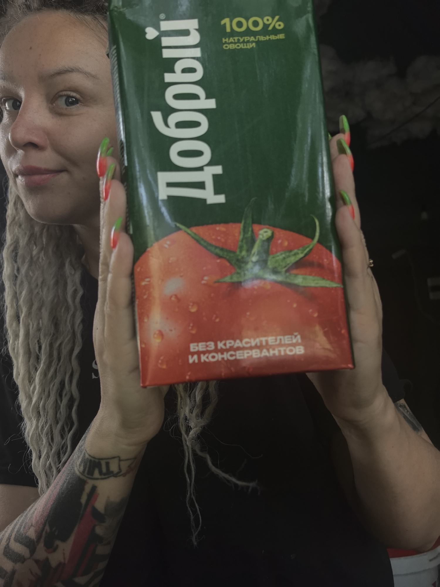 A woman with long blonde dreadlocks is holding a green and red carton of "Dobry" (Добрый) tomato juice. The carton is labeled "100% натуральные овощи" (100% Natural Vegetables) and "Без красителей и консервантов" (No Dyes or Preservatives). The woman's face is visible to the left of the carton, and her hands are holding the carton in front of her chest. She has red and green nail polish. The woman has tattoos on her left arm. The background is dark. The juice box is printed with a picture of a tomato. There is no indication from the image that the location is Noviy, Russia.