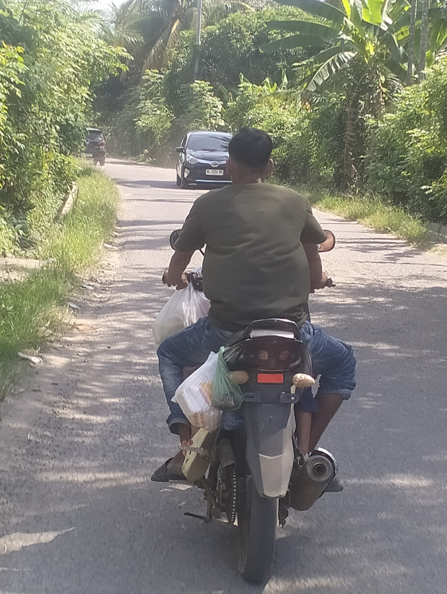 The image captures a street scene in Meunasahuket, Indonesia. A person is riding a motorcycle down a paved road lined with lush green vegetation and trees. The rider, wearing a green t-shirt and blue jeans, is carrying two plastic bags, one on either side of the motorcycle's rear. The bags appear to contain food items. In the background, a black car is visible further down the road, and the overall environment suggests a rural or semi-rural setting. The lighting indicates it is daytime, with sunlight casting shadows on the road.