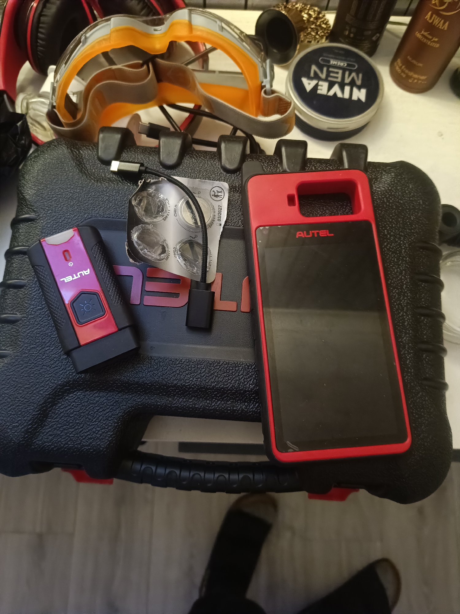 An overhead, close-up shot shows a black, textured, rectangular case resting on a light-colored, wooden floor. A red and black electronic diagnostic tool with a screen is placed on top of the case. To the left of the diagnostic tool is a smaller, red and black electronic device with a button and a light. A silver blister pack containing four circular items is also on the case, connected by a black wire to a USB adapter. In the background, several other items are visible including headphones, safety goggles, a small blue tub with "NIVEA MEN" printed on it, and a brown bottle with "AJWAA" on it. The items are arranged in a static, organized manner. The scene is indoors and does not provide any specific geographical indicators of Huntingdonshire, United Kingdom.