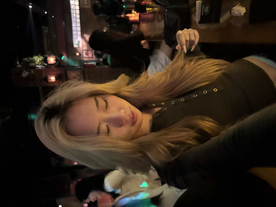 A young woman with blonde hair is resting her head, possibly asleep. She wears a black top with decorative buttons down the front. A person is visible behind her, holding her hair. The background is a dimly lit interior space that could be a bar or restaurant, with other patrons visible in the distance. The image is slightly tilted. A table with candles and other decorative items is also partially visible in the background.