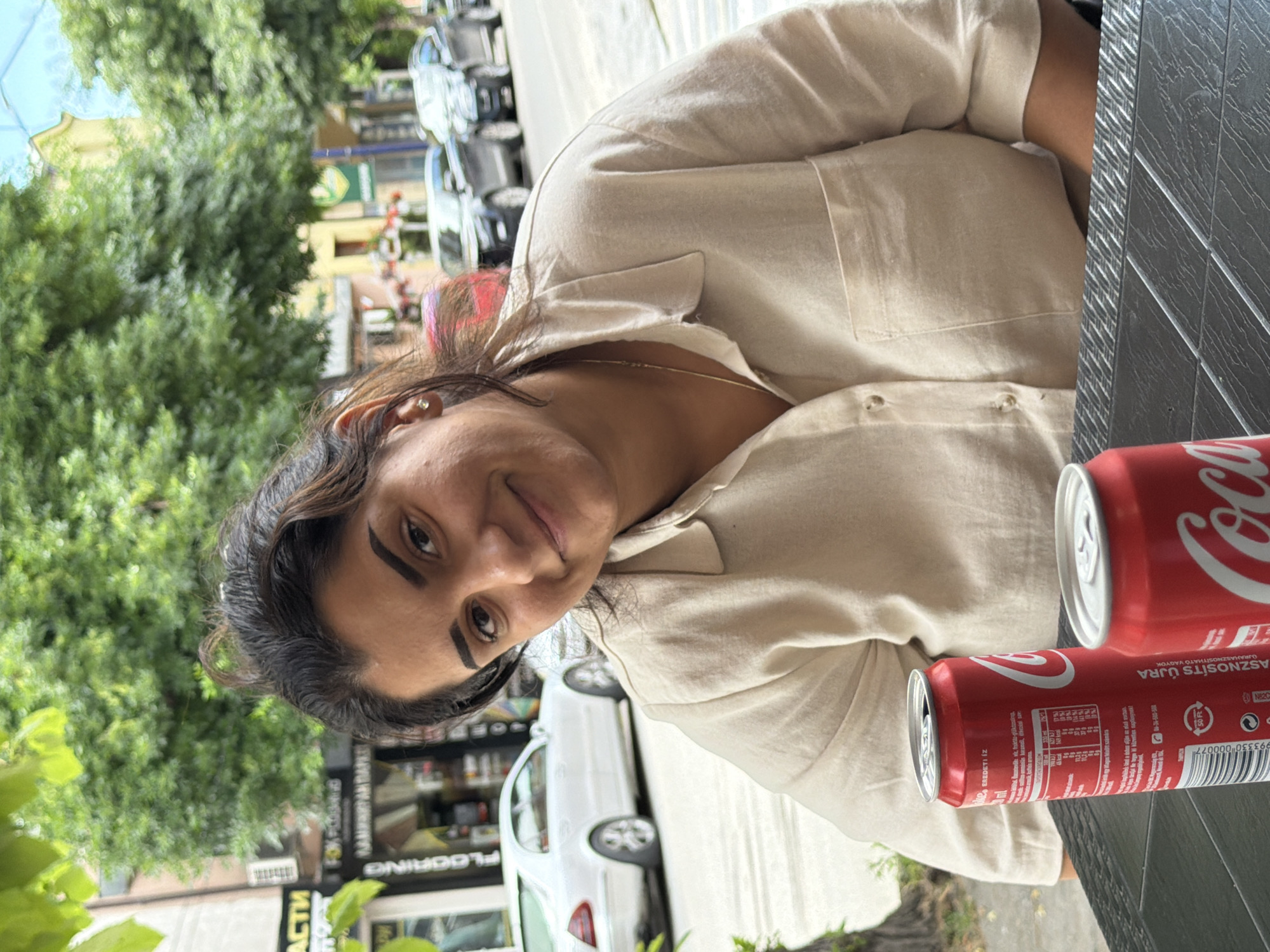 An image, rotated sideways, depicts a woman seated outdoors at a table, looking towards the viewer with a slight smile. She has dark, wavy hair and is wearing a light-colored, button-up collared shirt, possibly beige or off-white. A thin gold chain is visible around her neck, and she has a small, light-colored earring.
On the dark, textured table in the foreground, there are two red Coca-Cola cans. The closest can clearly displays the brand name "Coca-Cola" and Hungarian text such as "ASZNOSÍTS ÚJRA" (Recycle), "ÚJRAHASZNOSÍTHATÓ VAGYOK" (I am recyclable), and "EREDETI ÍZ" (Original Taste).
The background suggests a vibrant urban setting during daylight hours, likely in Pazardzhik, Bulgaria, as indicated by the context. Lush green trees line a street where white cars are parked. Buildings are visible behind the trees and vehicles. Partially visible, rotated text on a building sign includes "FLOORING", "ЛАМИНИРАН ПАРК" (Laminated Flooring), and "АСТИ" (ASTI), confirming the Bulgarian context. Another sign seems to show "МАГАЗИН" (Shop). The lighting is bright, indicating good weather. The overall scene conveys a casual moment at an outdoor cafe or restaurant.