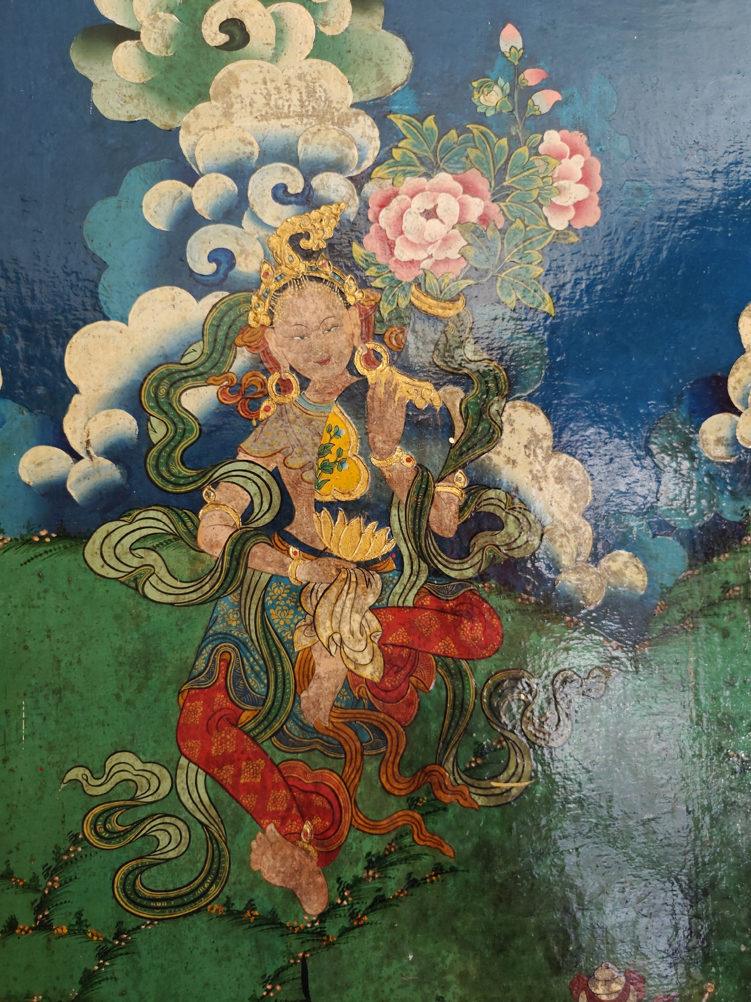 The image depicts a painted figure, possibly a deity or celestial being, in a traditional style. The figure is adorned with golden jewelry and flowing green serpentine strands framing the body. In the upper right corner of the image are pink flowers. The figure is positioned against a backdrop of green and blue, with depictions of swirling clouds. This image does not contain sufficient data to provide information about the location in the city of Fatehpur, India.