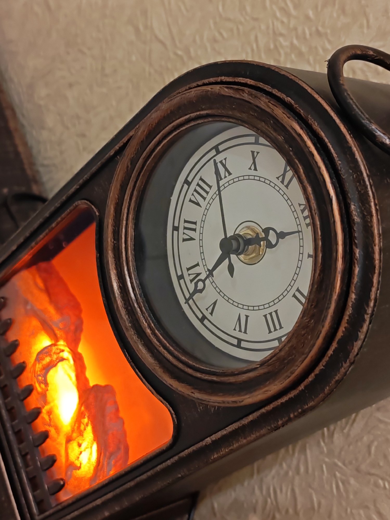The image shows a close-up of a decorative clock with a Roman numeral face. The clock is designed to look like a miniature fireplace, with a faux fire glowing behind a glass panel on the left side of the image. The clock itself is made of dark metal with a bronze-colored trim around the face. The hands of the clock are pointing to approximately 2:50. The background is a textured wall. There are no people or animals visible in the image. The location is not specified in the image.