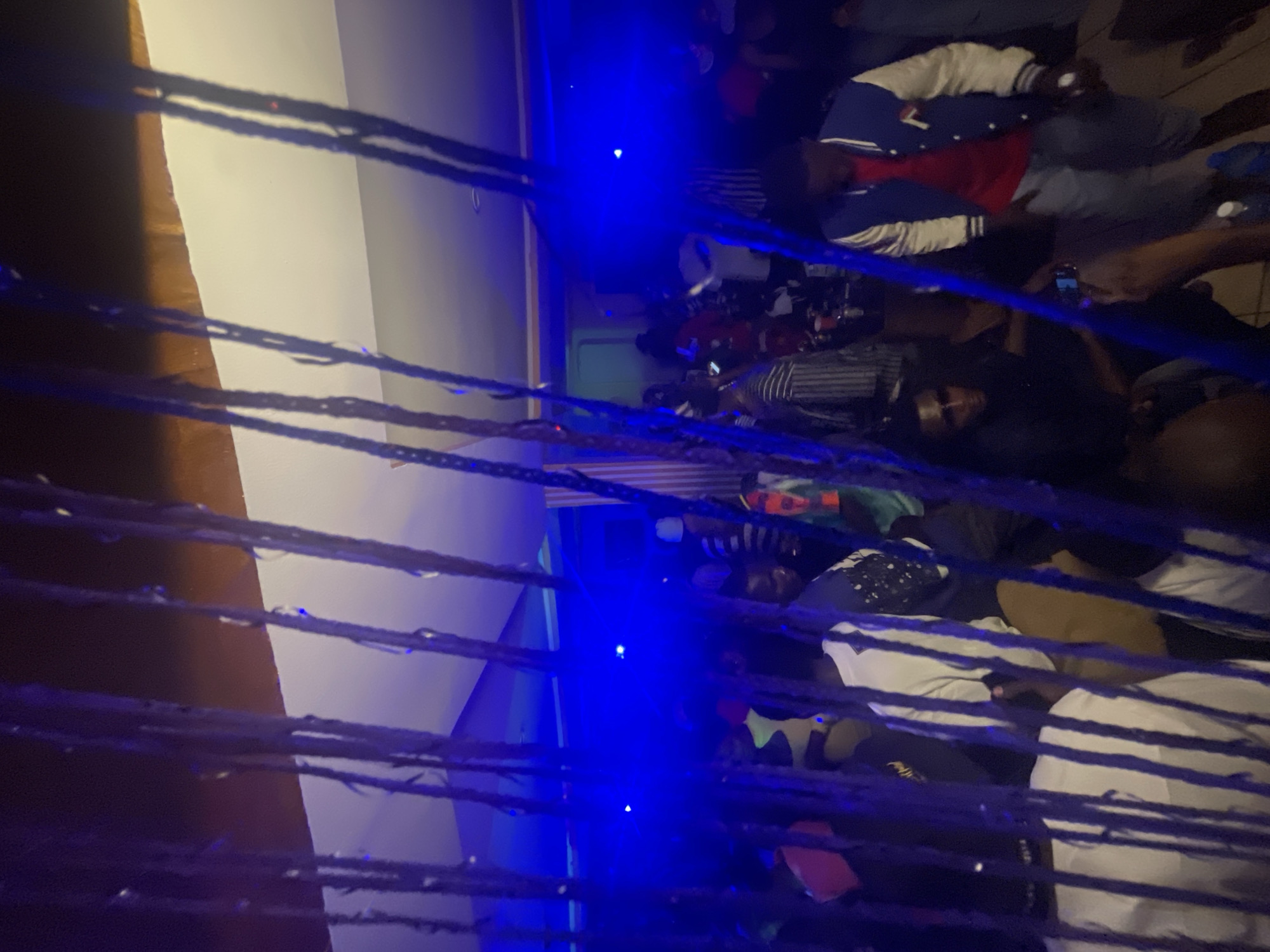 The image captures a dimly lit indoor scene, likely a party or gathering, illuminated by vibrant blue lights. The foreground is partially obscured by dark, rope-like strands, possibly part of a decorative element or curtain. Beyond these, a crowd of people is visible, many in casual attire.
In the mid-ground, several individuals are distinguishable. To the left, a person in a white t-shirt is partially visible. Further to the right, a person wearing a blue and white varsity jacket over a red shirt stands, with grey pants. Another individual in the mid-ground wears sunglasses and a black top. The background shows more indistinct figures and the general ambiance of a lively event, with some people dancing or socializing. The ceiling is visible at the top, featuring a combination of white and wooden elements. The overall impression is one of a social event in a somewhat confined space, characterized by low lighting and a festive atmosphere. The location in Wumat, Nigeria, suggests a cultural context for this gathering.