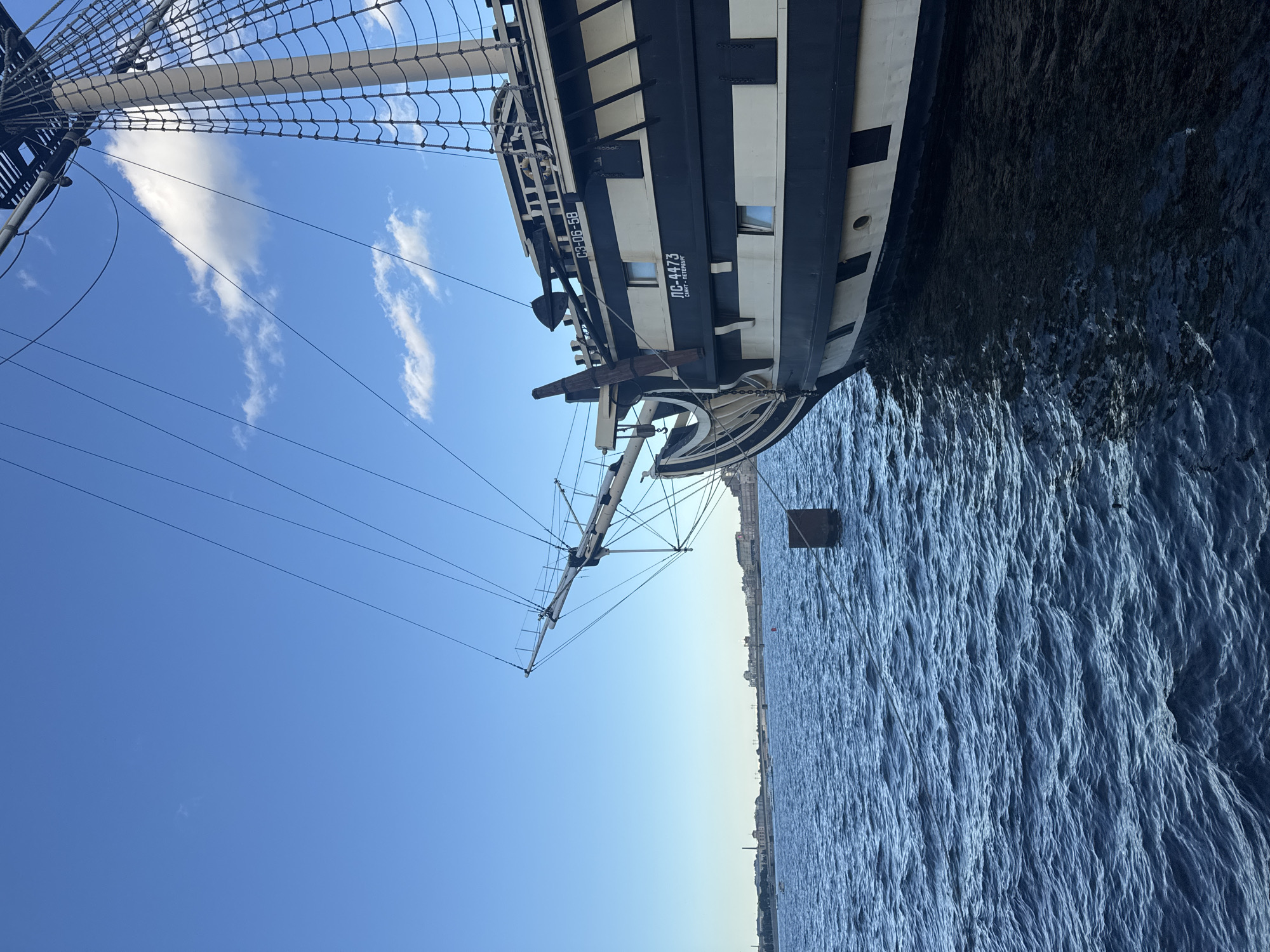 The image captures a clear, sunny day in Saint Petersburg, Russia, with a portion of a historic sailing ship prominently featured in the foreground. The ship, painted black and white, has multiple masts and rigging, suggesting it is a replica or preserved vessel. The bow of the ship is angled towards the viewer, with visible hull details and windows.
The ship is docked in the Neva River, as indicated by the choppy blue water reflecting the sky. In the background, the distant skyline of Saint Petersburg is visible, featuring a bridge and various buildings. The sky is a vibrant blue with a few wispy clouds.
The scene appears to be a tourist attraction or a historical exhibit, given the nature of the ship. The lighting suggests it's likely late morning or early afternoon. No people are visible in the frame. The overall impression is one of maritime history set against the backdrop of a modern city.