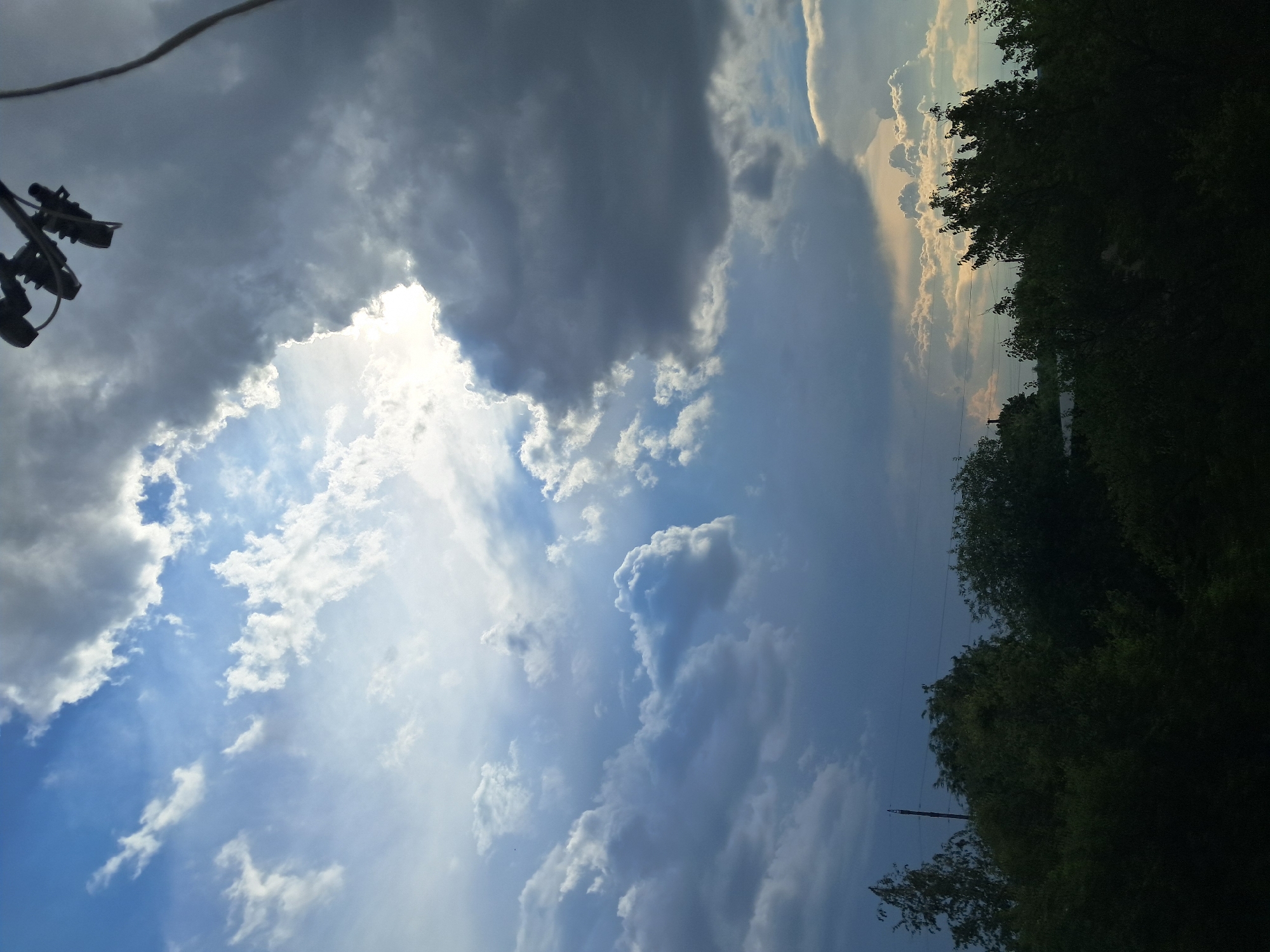The image depicts a dramatic sky over Smila, Ukraine, with a prominent dark storm cloud partially obscuring the sun. Rays of light break through the clouds, creating a contrast between light and shadow. Below the sky, a dense line of dark green trees forms the foreground, with hints of a few structures and power lines visible amidst the foliage. The overall impression is one of an approaching or receding storm, with the landscape cast in a somewhat somber light.