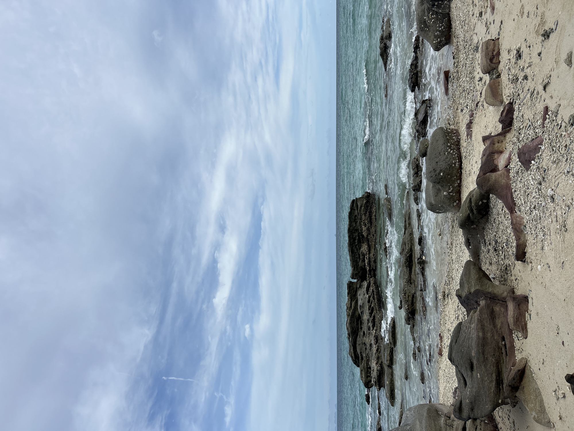 This is a landscape photograph of a rocky beach under a cloudy sky. The foreground consists of sand and various sizes of rocks and pebbles. Some of the rocks are large and dark, while others are smaller and reddish-brown. The middle ground features the ocean with gentle waves breaking against a cluster of larger, dark rocks in the water. The sea is a turquoise color. The background is dominated by a pale blue sky with streaks of white clouds and a distant, hazy landmass or islands on the horizon. The location is described as Phru Nai, Thailand. There are no people or any explicit human-made objects visible in the scene.