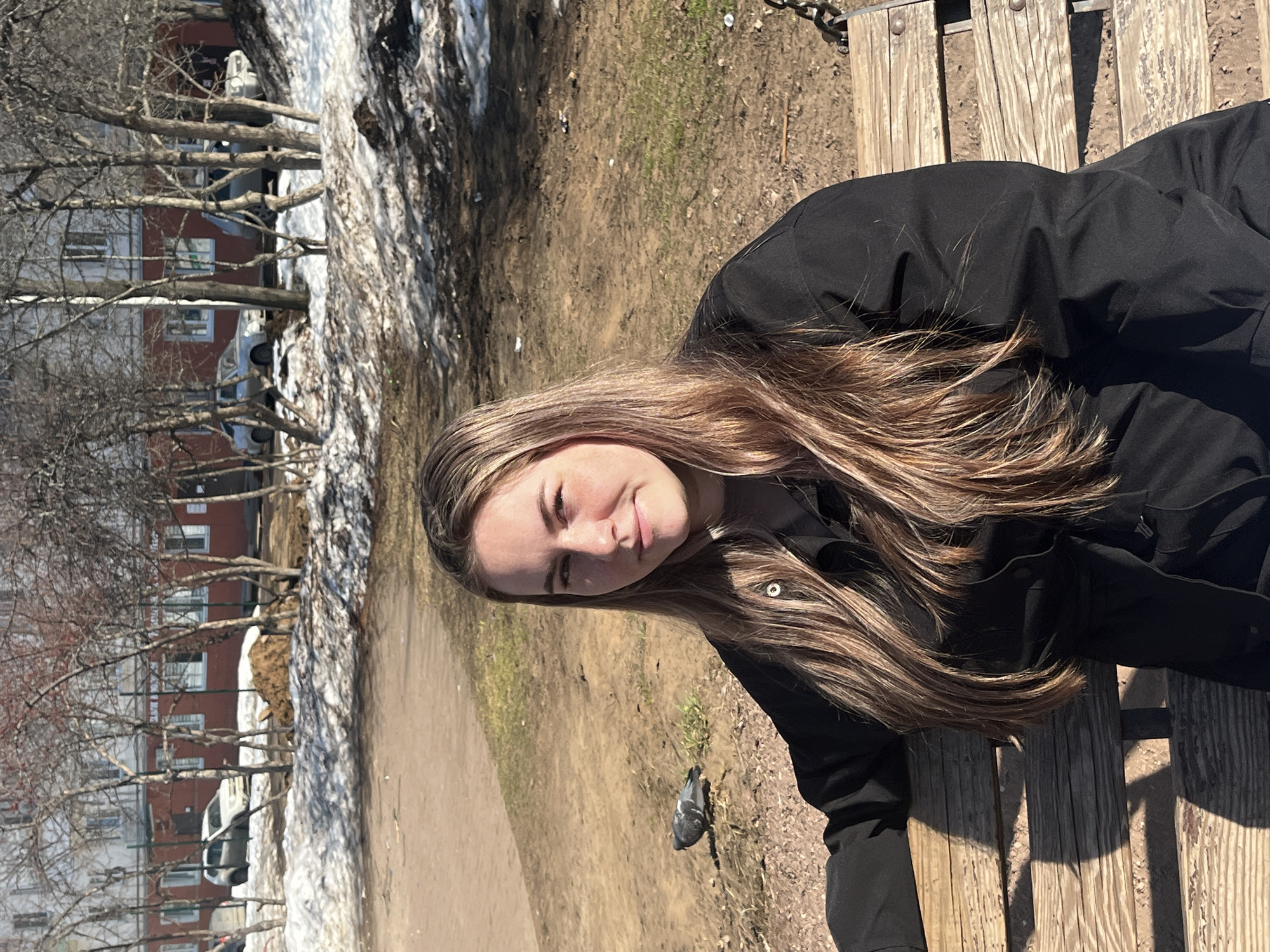 A woman with long brown hair is lying on a wooden park bench in Chaikovsky, Russia. The background shows an urban park with melting snow, bare trees, and a path, and a multi-story residential building. A pigeon is on the ground nearby. The woman is wearing a black jacket.