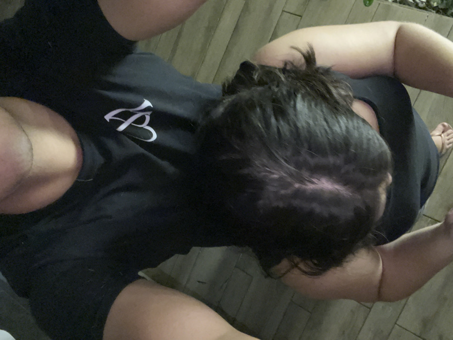 A high-angle photograph captures two individuals in what appears to be an indoor or covered outdoor setting with light-colored, possibly tiled or planked flooring. The person holding the camera is partially visible from the mid-chest upward, wearing a black short-sleeved t-shirt adorned with a white stylized 'M' or similar symbol on the left chest, and a thin chain around their neck. Positioned directly below and in front of the camera-holder is a second individual, viewed from the top of the head to the upper torso. This person has dark, wavy hair with a prominent part revealing some scalp, and wears a dark-colored tank top or dress. Their right foot, wearing a light-colored flip-flop, is visible on the floor. A small patch of green foliage is present on the floor's far right edge. No visual identifiers within the image confirm the location as Coatzacoalcos, Mexico.