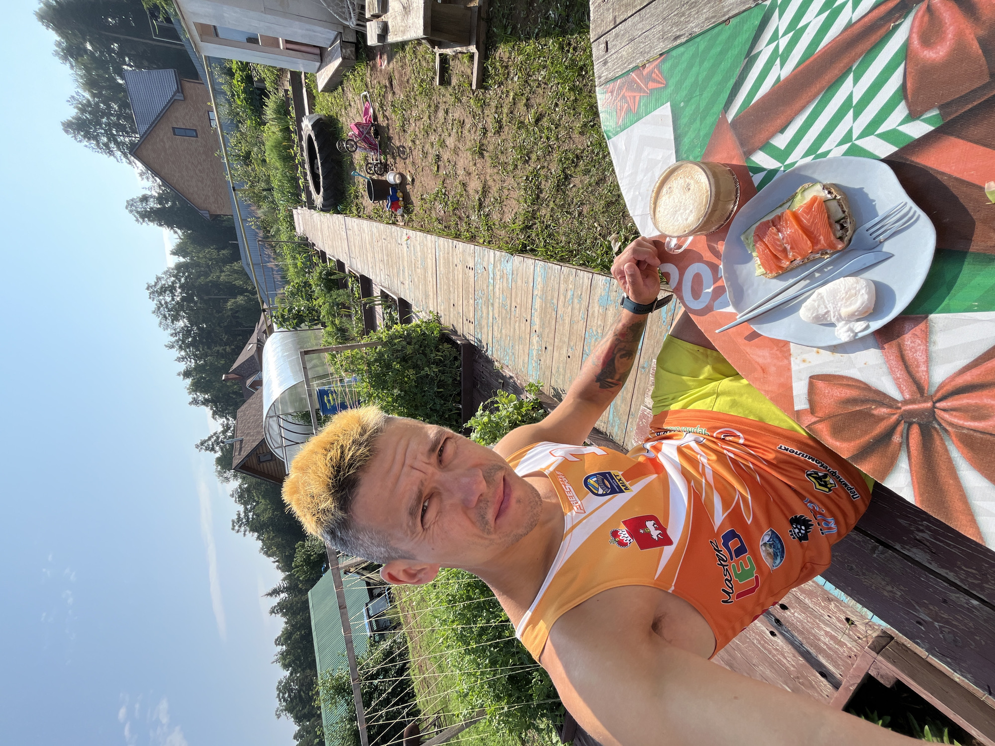A man with dyed blond hair sits at a wooden table outdoors, taking a selfie. He is wearing an orange tank top with various logos and yellow shorts. On the table in front of him is a plate with a slice of bread topped with salmon and a poached egg, and a cup of coffee. The background shows a garden with raised beds, a greenhouse, trees, and houses. The location is identified as Chaikovsky, Russia.