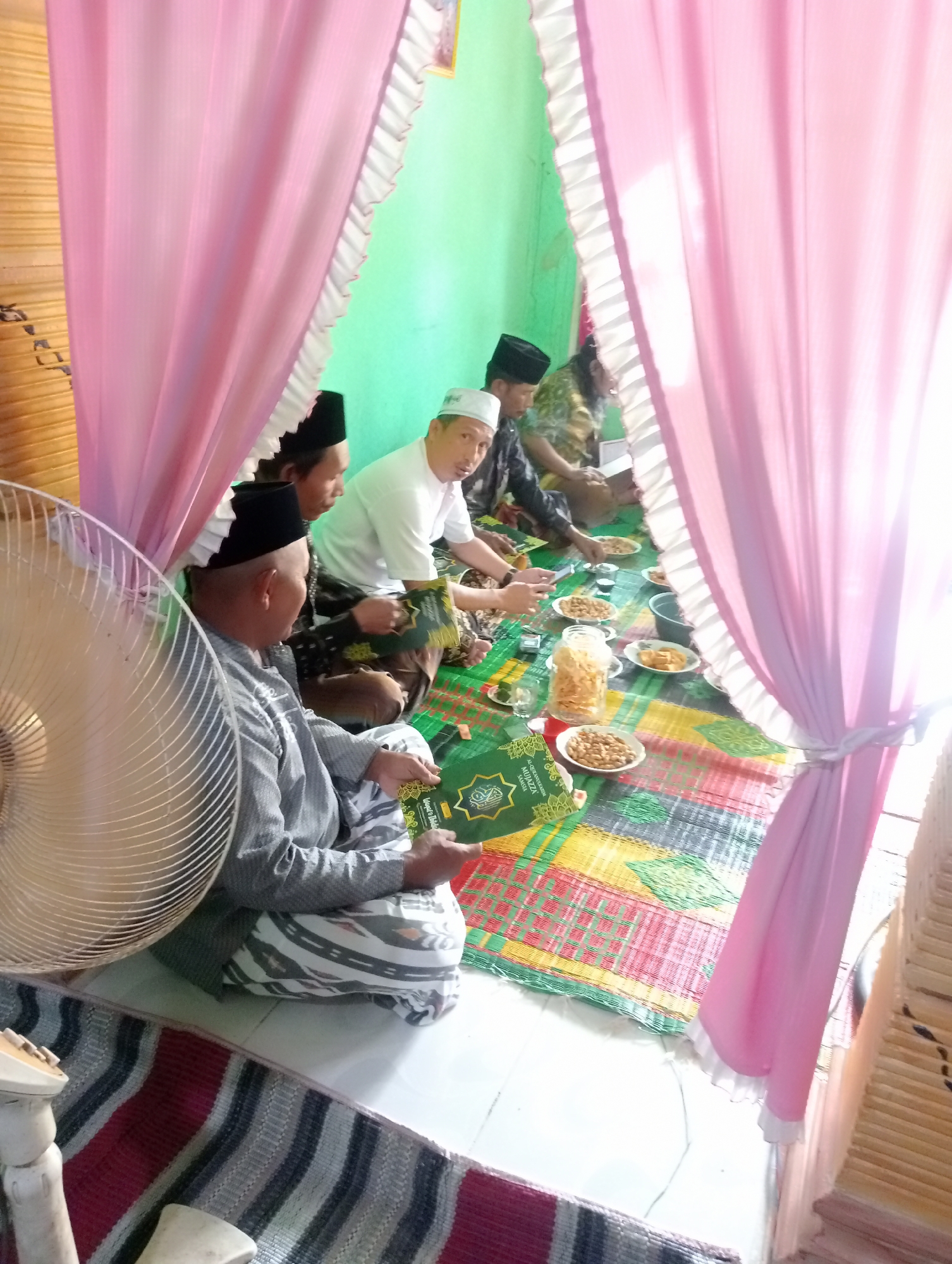 The image depicts a gathering of at least six men seated on the floor on a woven mat, in what appears to be a room in Kota Negara Timur, Indonesia. The men are dressed in traditional Islamic attire, including peci caps and shirts. Some are wearing sarongs. The room is decorated with pink curtains that frame the scene. In the foreground, a large fan is visible. On the mat in front of the men, plates of food and a jar of snacks are laid out, suggesting a communal meal or event. One man is holding a green book with Arabic script. The background wall is painted a light green color. The lighting suggests it is daytime, possibly with sunlight streaming in from the right.