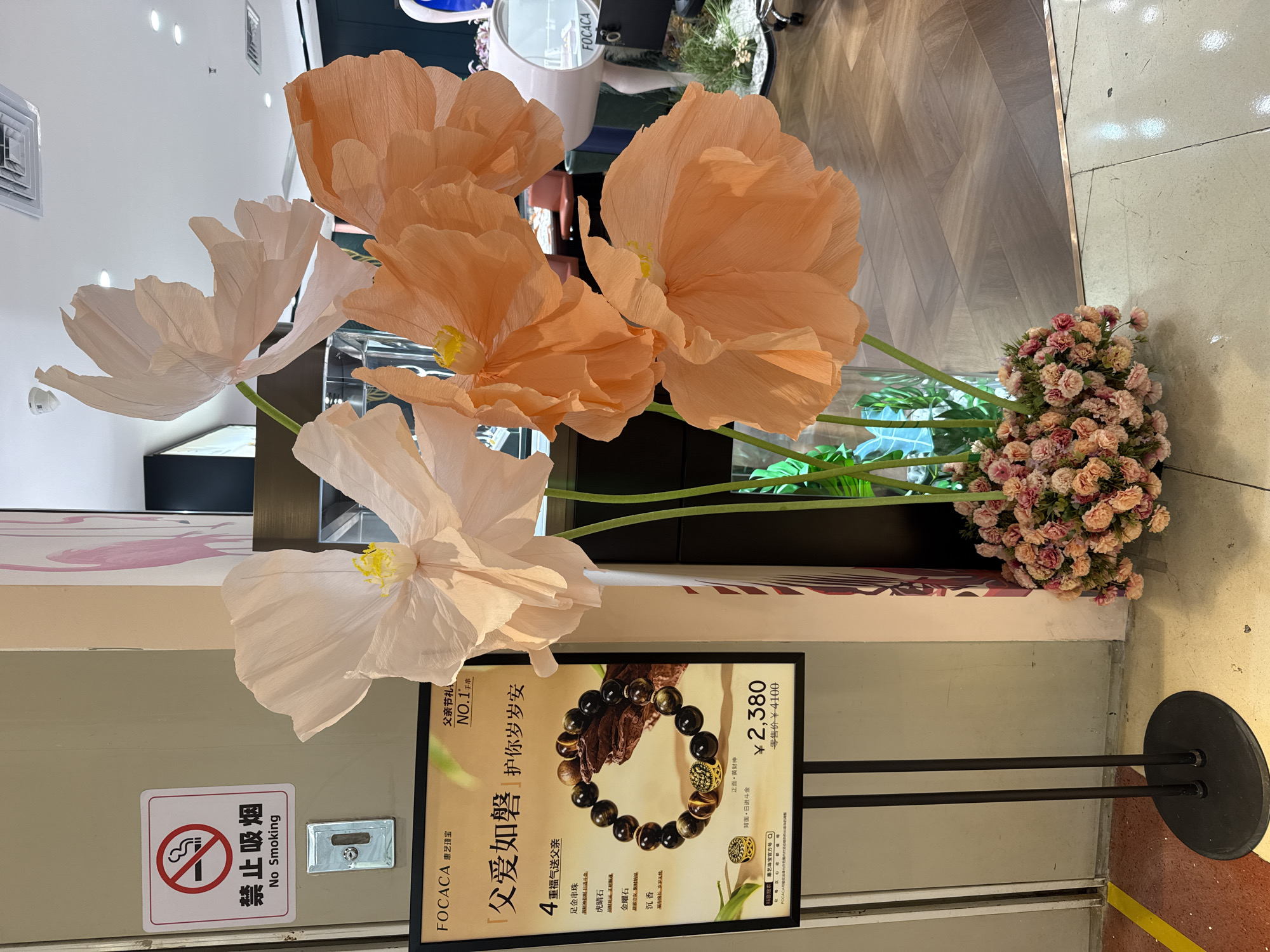 A no smoking sign is mounted on a neutral-colored wall, to the left of a display of large, artificial flowers. The sign is white with a red border and features a cigarette with a red line through it, and the text "禁止吸烟" and "No Smoking" below. Below the sign is a black sign holder with a digital display reading "¥2,380" and a crossed-out price of "¥4,100". The flowers, which appear to be made of paper or fabric, are in shades of pale pink and peach and are arranged in a cluster of smaller pink flowers at their base. They are positioned in front of a display of jewelry. The floor is a combination of light-colored tiles and wood flooring. The scene appears to be in a commercial establishment, possibly a jewelry store, in Huizhou, China.