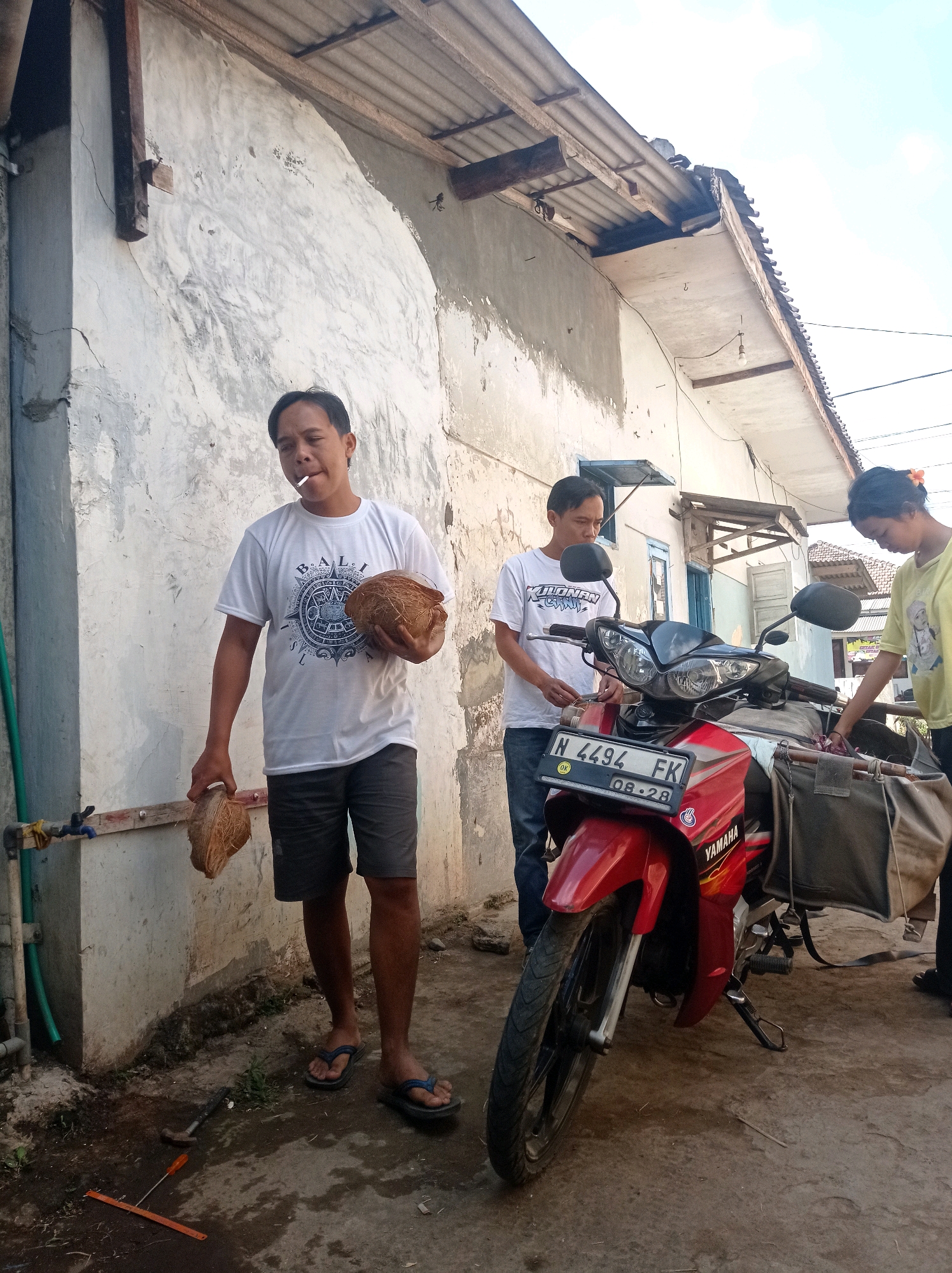 In Purwosekar, Indonesia, three individuals are present in an outdoor setting beside a red Yamaha motorcycle parked on a concrete surface. A man in the foreground, wearing a white t-shirt and grey shorts, holds two halves of a coconut. He is smoking a cigarette. Behind him, another man in a white t-shirt and blue jeans stands near the motorcycle, appearing to interact with it. To the right, a woman in a yellow t-shirt is also near the motorcycle, possibly attending to it. The background features a weathered concrete wall and a corrugated metal roof, suggesting a modest dwelling or structure. A green hose is visible on the left side of the image, connected to a faucet. Tools, including a hammer and a ruler-like object, are scattered on the ground near the man in the foreground.