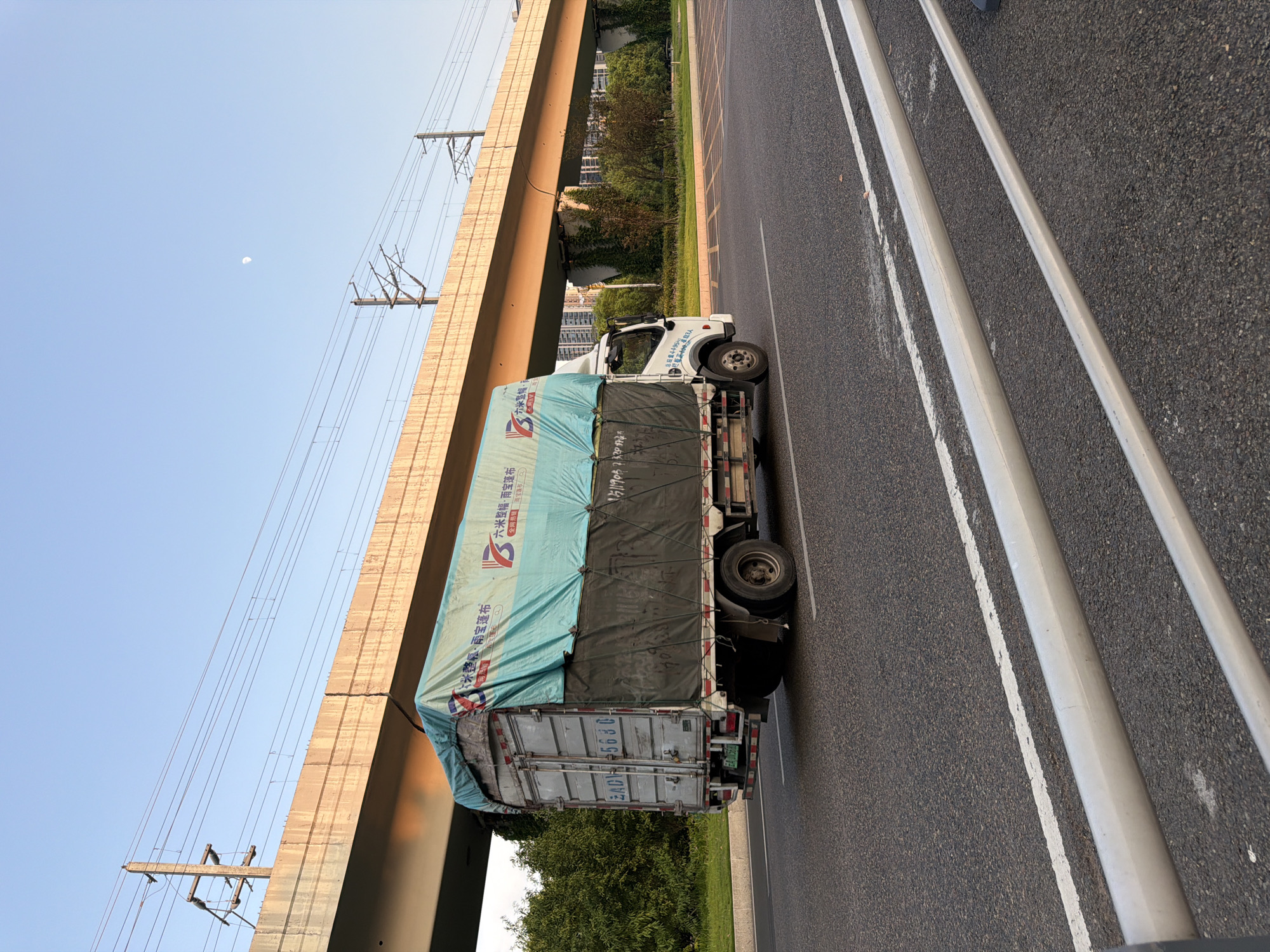A white box truck with a blue tarp covering its cargo is driving on a paved road. The truck has license plate "EA 15G8". The truck is under an elevated concrete structure, likely a highway overpass, with overhead power lines. Trees and buildings are visible in the background. The sky is clear and blue, with a sliver of the moon visible. The scene appears to be in a semi-urban or suburban area in Jiangjin, China.