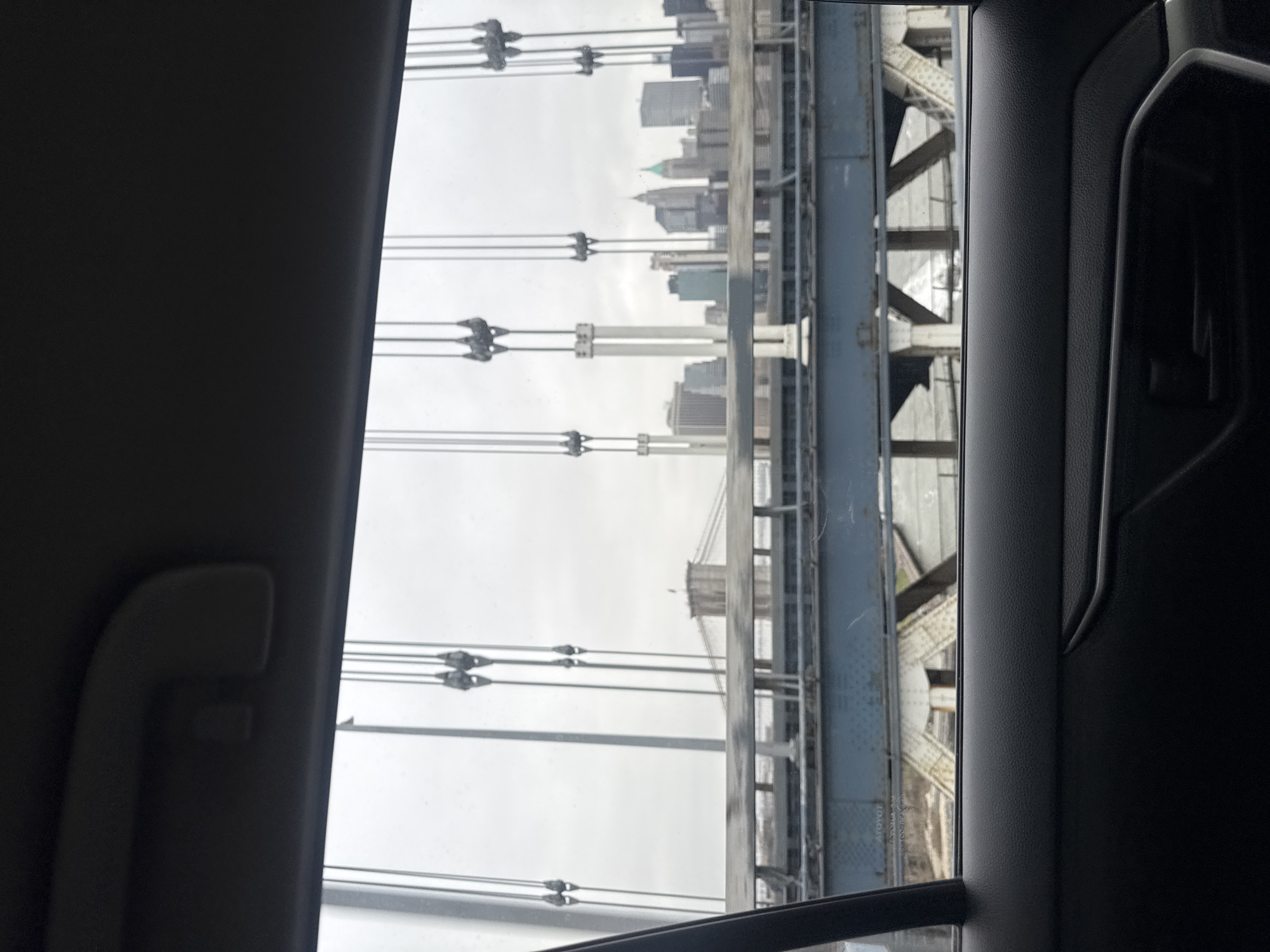 The image shows a view from inside a car, looking out through the side window. The subject is the Manhattan Bridge, a suspension bridge in New York City, USA. The bridge's steel structure and suspension cables are prominently displayed. In the background, the skyline of lower Manhattan is visible in the distance, with several skyscrapers discernible. The sky is overcast. The interior of the car is visible in the foreground, including the door panel, window frame, and handle.
