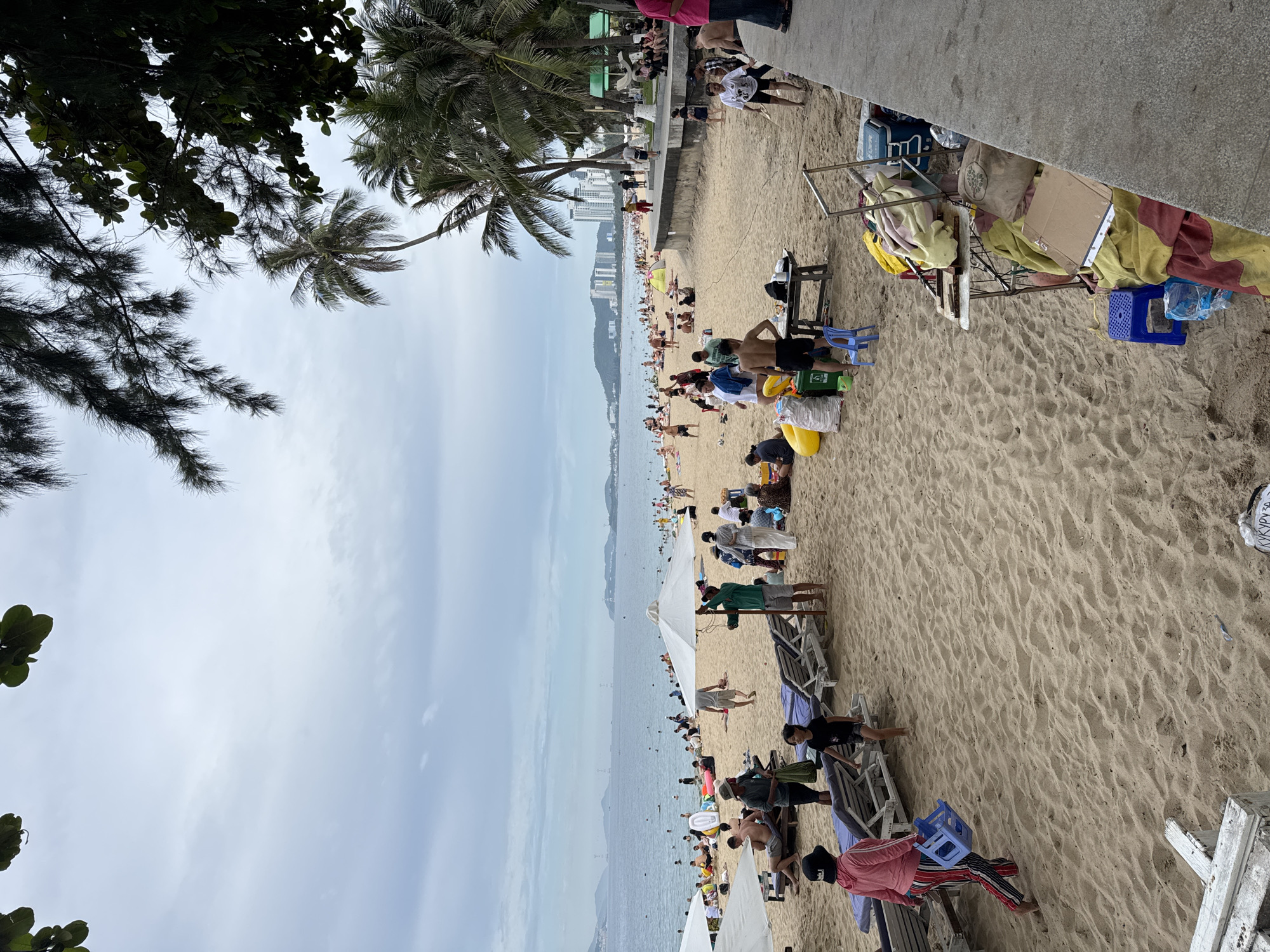 The image depicts a busy beach scene in Nha Trang, Vietnam. The sandy beach is populated with numerous individuals, some lounging on beach chairs, others walking or engaging in various activities. In the background, the ocean is visible, with several boats scattered across its surface. Along the coastline, modern buildings and palm trees suggest an urban environment. Several beach umbrellas provide shade, and various objects such as coolers, bags, and chairs are scattered across the sand, indicating a typical day at a popular tourist destination. The sky is overcast, suggesting potentially mild weather.