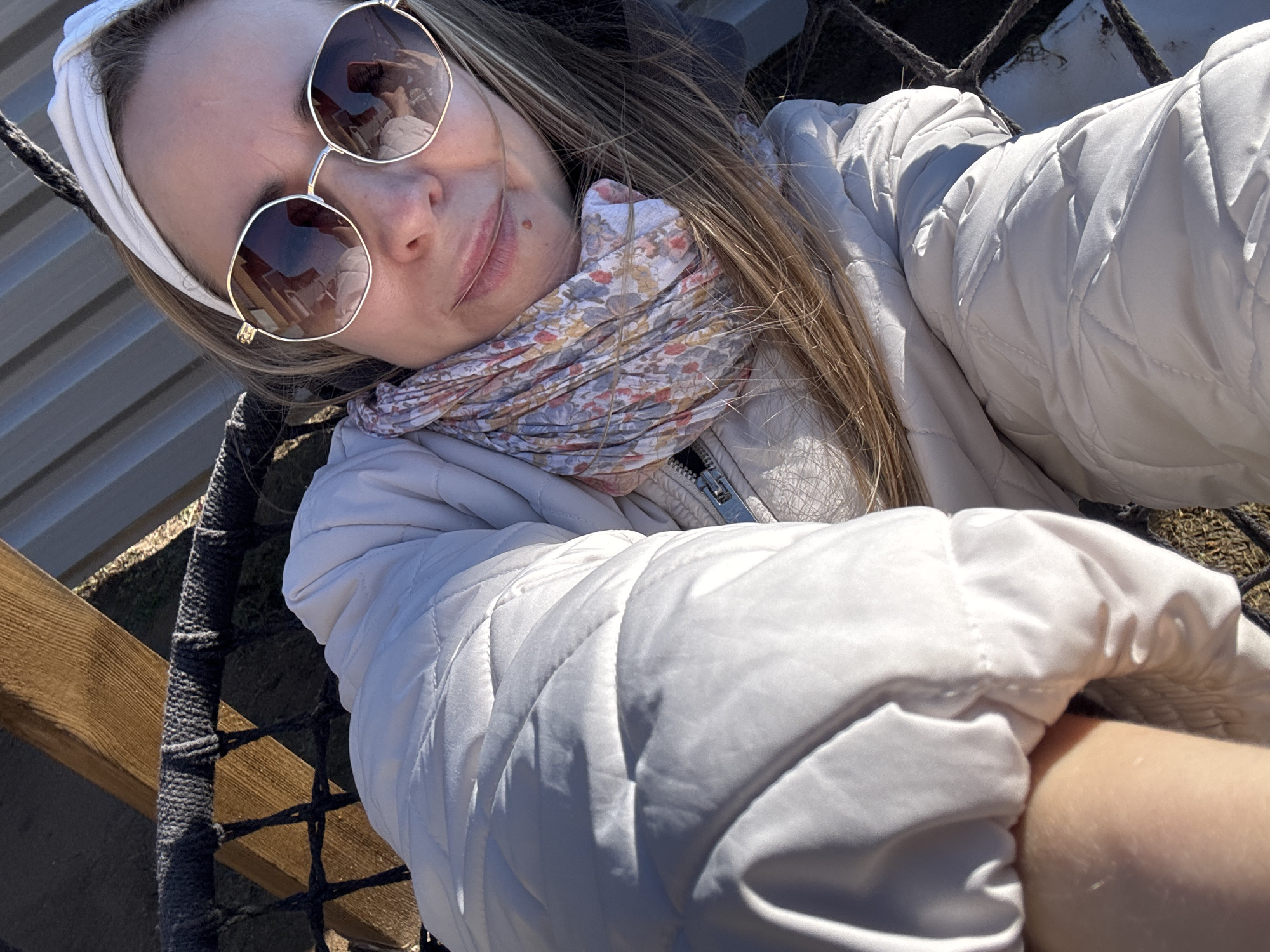 A person, presumably female, is taking a selfie. The individual is wearing sunglasses, a white headband, a patterned scarf, and a white quilted jacket. The background reveals a wooden structure and a rope net. The location is presumed to be Chaikovsky, Russia.