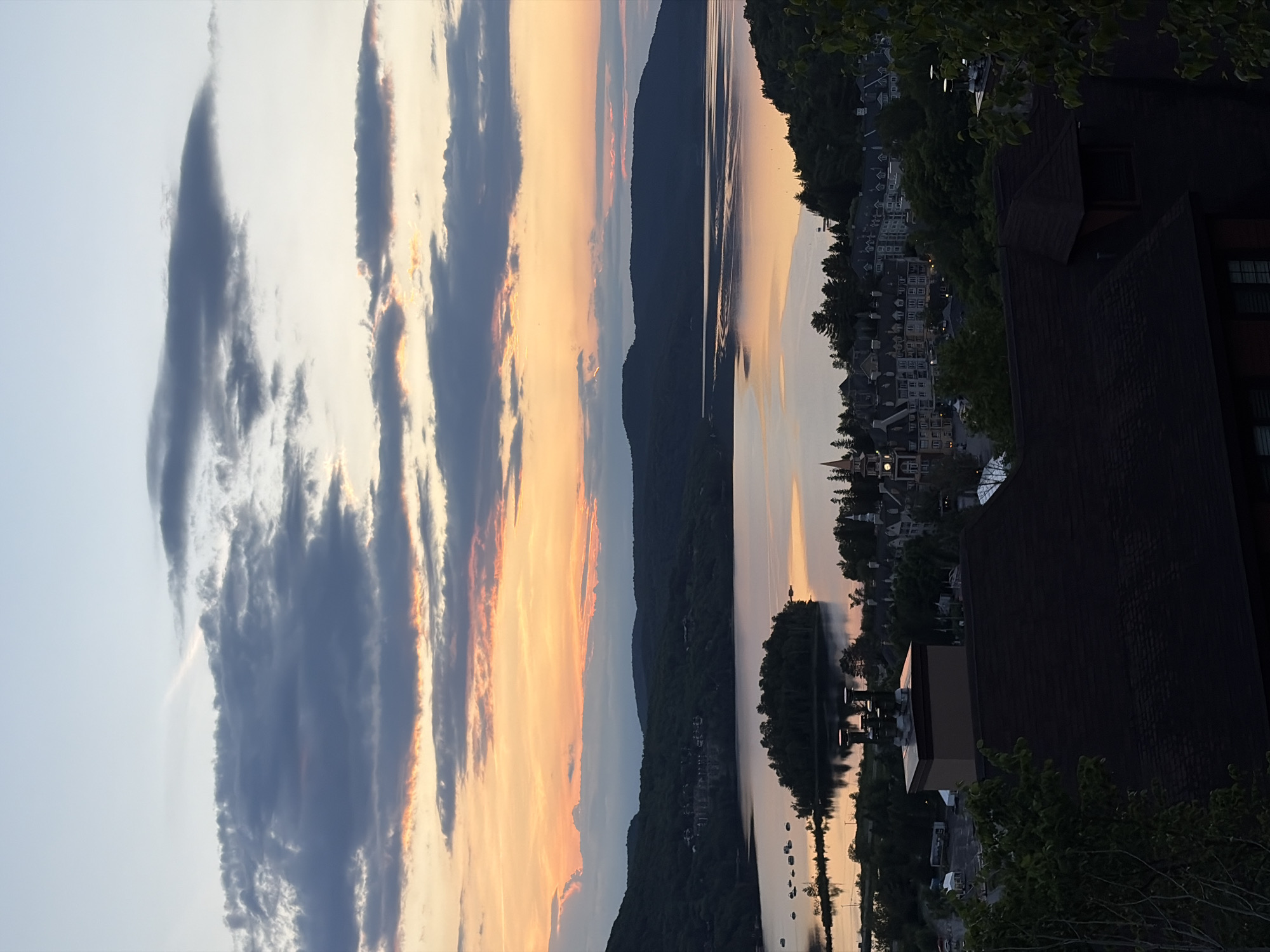 The image depicts a scenic view of Mont-Tremblant, Canada, at dusk. In the foreground, a dark-shingled roof of a building dominates the lower portion of the frame. Above this, a cluster of metallic chimneys stands out against the twilight sky.
The midground reveals a charming village nestled among lush green trees, characterized by its European-style architecture with numerous gabled roofs and dormer windows. A prominent clock tower with a spire marks the center of the village. Beyond the village, a tranquil body of water, likely a lake, reflects the colors of the sunset. A small, tree-covered island sits in the middle of the lake.
The background features rolling hills covered in dark green foliage, receding into the distance. The sky above is a spectacle of color, with hues of orange, pink, and yellow from the setting sun blending into shades of gray and blue from the clouds. The overall impression is one of peacefulness and natural beauty at the end of the day in this picturesque Canadian location.