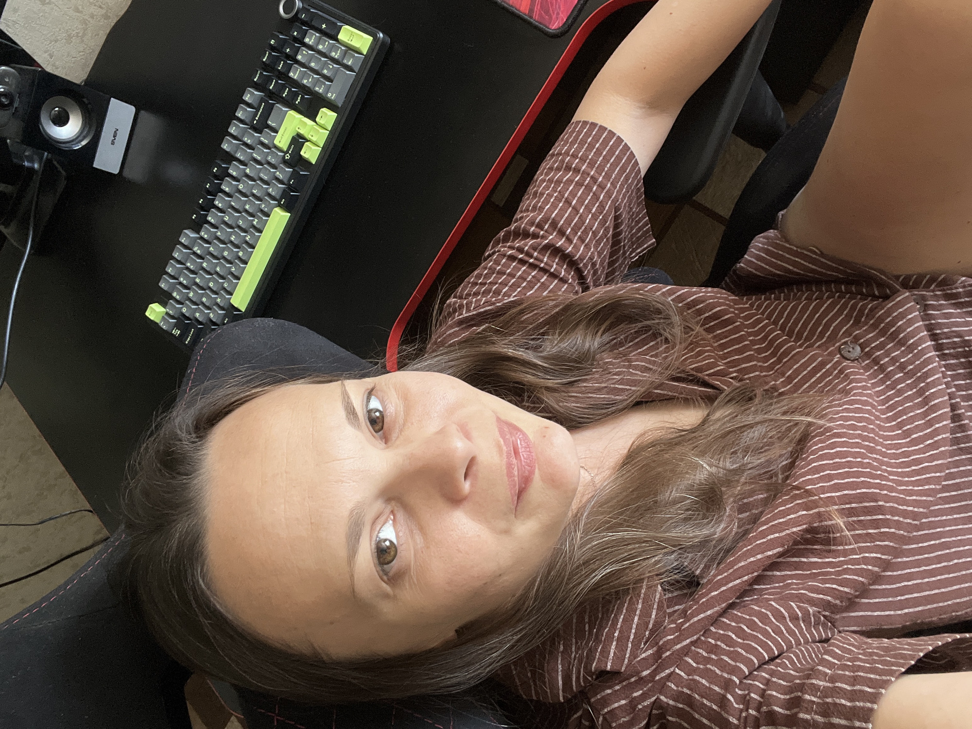 A woman is seated in a black gaming chair, facing forward and looking directly at the camera. She has long, brown hair and is wearing a brown, pinstriped shirt. Her arms are resting on a black desk that is positioned in front of her. On the desk, there is a black keyboard with lime green accents and a silver speaker. The background shows a portion of the chair and the desk, suggesting an indoor setting. The image appears to be taken from a slightly elevated angle. The scene is likely her personal workspace or gaming setup.