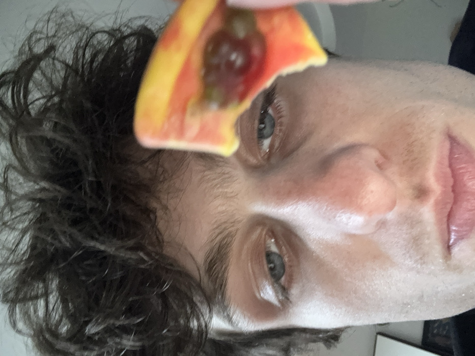 A close-up, eye-level selfie-style photograph depicts a male individual holding a piece of fruit near his face. The man has curly, dark brown hair that appears disheveled, blue eyes with dark brows, and visible stubble on his lower face. His skin tone is light, with some reddish areas around the nose and cheeks. He looks directly towards the camera with a neutral expression. A bright orange and yellow piece of fruit, possibly a slice of papaya or mango, is held in the foreground, partially obscuring the upper right portion of his face, including his right eye and forehead. The fruit shows an exposed interior with dark, possibly seed-filled, red pulp. The background is out of focus and light-colored, suggesting an indoor setting, with a faint, dark rectangular shape visible in the lower left corner, possibly a framed object or mirror. The specific location within the city of Vincennes, France, cannot be determined from the visual information provided.