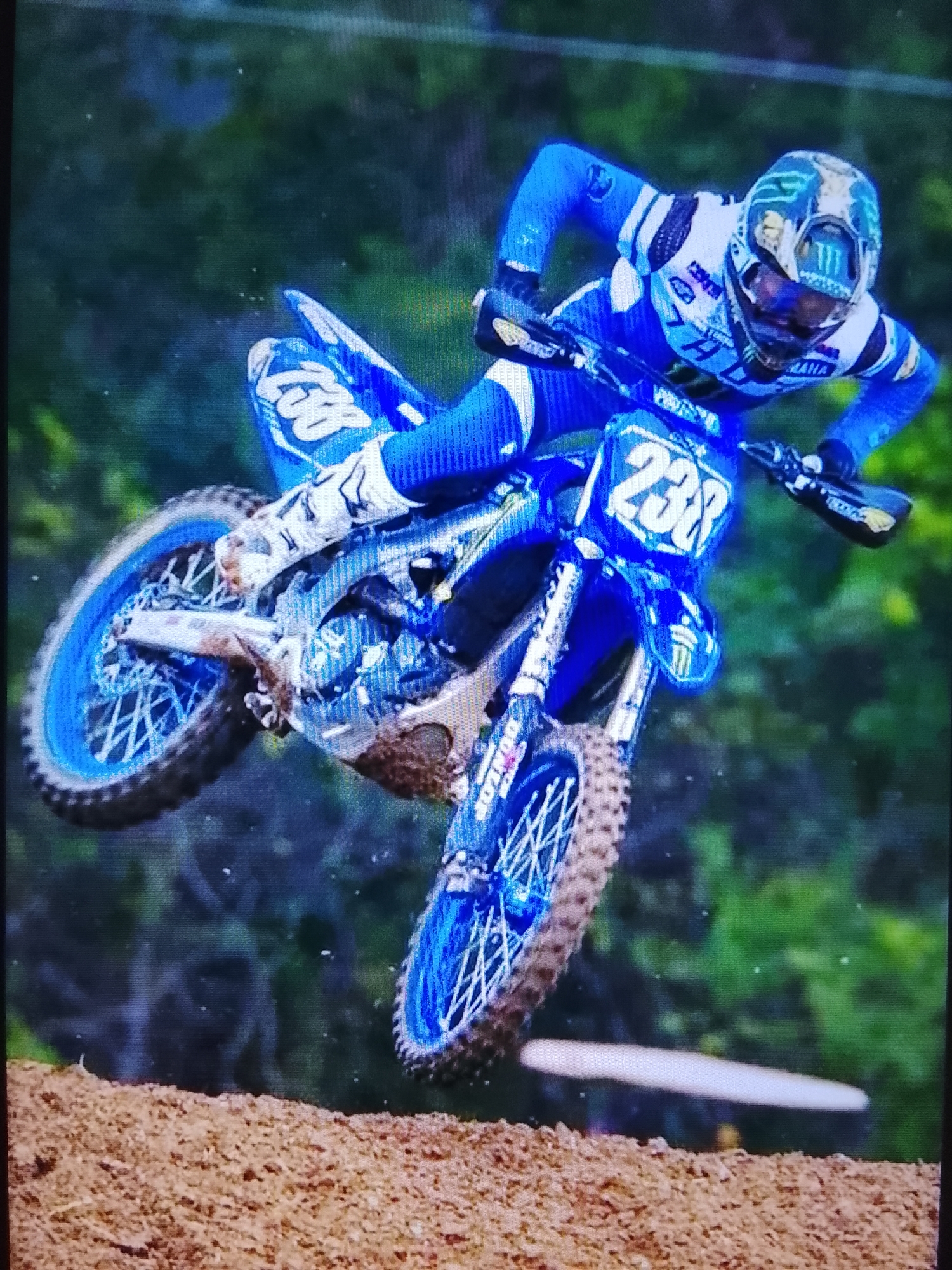A motocross rider in a blue and white suit with a blue helmet is captured mid-air on a blue and white dirt bike. The rider is leaning forward, intensely focused, with the bike angled slightly. The front wheel is turned to the left, and both wheels appear to be off the ground. The bike has the number "238" prominently displayed on a blue plate on the front. Mud is splattered on the bike's frame and wheels, suggesting recent activity on a dirt track.
The background consists of blurred green foliage, indicative of an outdoor setting, likely a motocross track surrounded by trees. The ground in the foreground is a rough, brown dirt surface. The lighting suggests it is daytime, with natural light illuminating the scene. The image captures a dynamic moment of action and athleticism in a motocross event. Visible text includes "YAMAHA" on the bike and the rider's suit, "DUNLOP" on the tire, and a "Monster Energy" logo on the helmet.