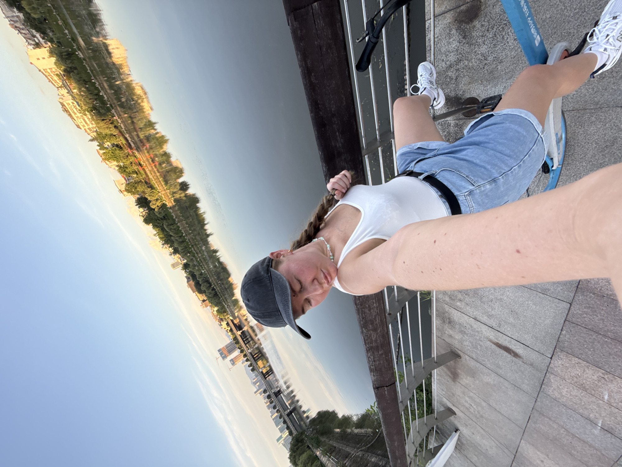 A woman is taking a selfie on a blue bicycle on a pathway next to a body of water in Yiwu, China. She is wearing a white tank top, denim shorts, a black baseball cap, and white sneakers. Her hair is in a braid. The background shows a city skyline reflected in the water, with buildings and trees visible. The sky is clear and blue. The pathway is paved with large gray stones. A metal railing runs along the edge of the pathway. The water appears calm and reflects the sky and the city. The overall scene suggests a leisurely activity in an urban environment.