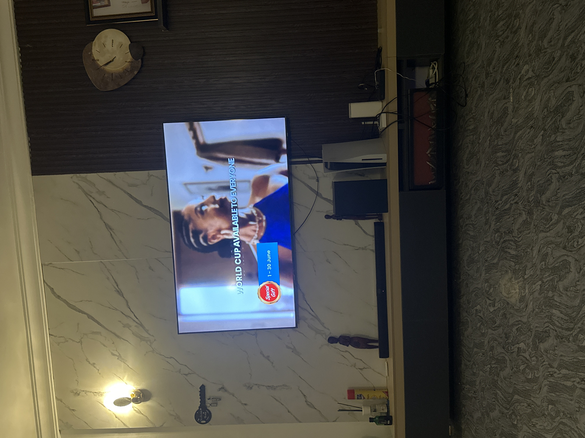 This image depicts the interior of a modern living room, likely in Gwarinpa, Nigeria. A large flat-screen television is mounted on a wall, which is a mix of marble-patterned and dark wood-paneled sections. The television displays a graphic that reads "WORLD CUP AVAILABLE TO EVERYONE" with a "Special Gift" banner and a date range of "1 - 30 June".
On a long, dark gray media console below the TV, there are several items including a soundbar, a black game console (likely an Xbox), a white PlayStation 5, a white cylindrical object, and some black cables. To the left of the console, on a wooden shelf, are various bottles and containers, including what appears to be a yellow spray can and a green bottle. A small wooden figurine is placed on the shelf as well.
To the right of the television, on the wood-paneled wall, a decorative clock with an abstract design is mounted. Above the clock, a framed picture is visible. A wall-mounted light fixture with a warm glow is on the left side of the marble-patterned wall. A decorative metal key holder with the words "Welcome Home" is also mounted on the wall near the light fixture. The floor has a patterned, possibly marbled, design. The overall impression is of a well-decorated and contemporary living space.