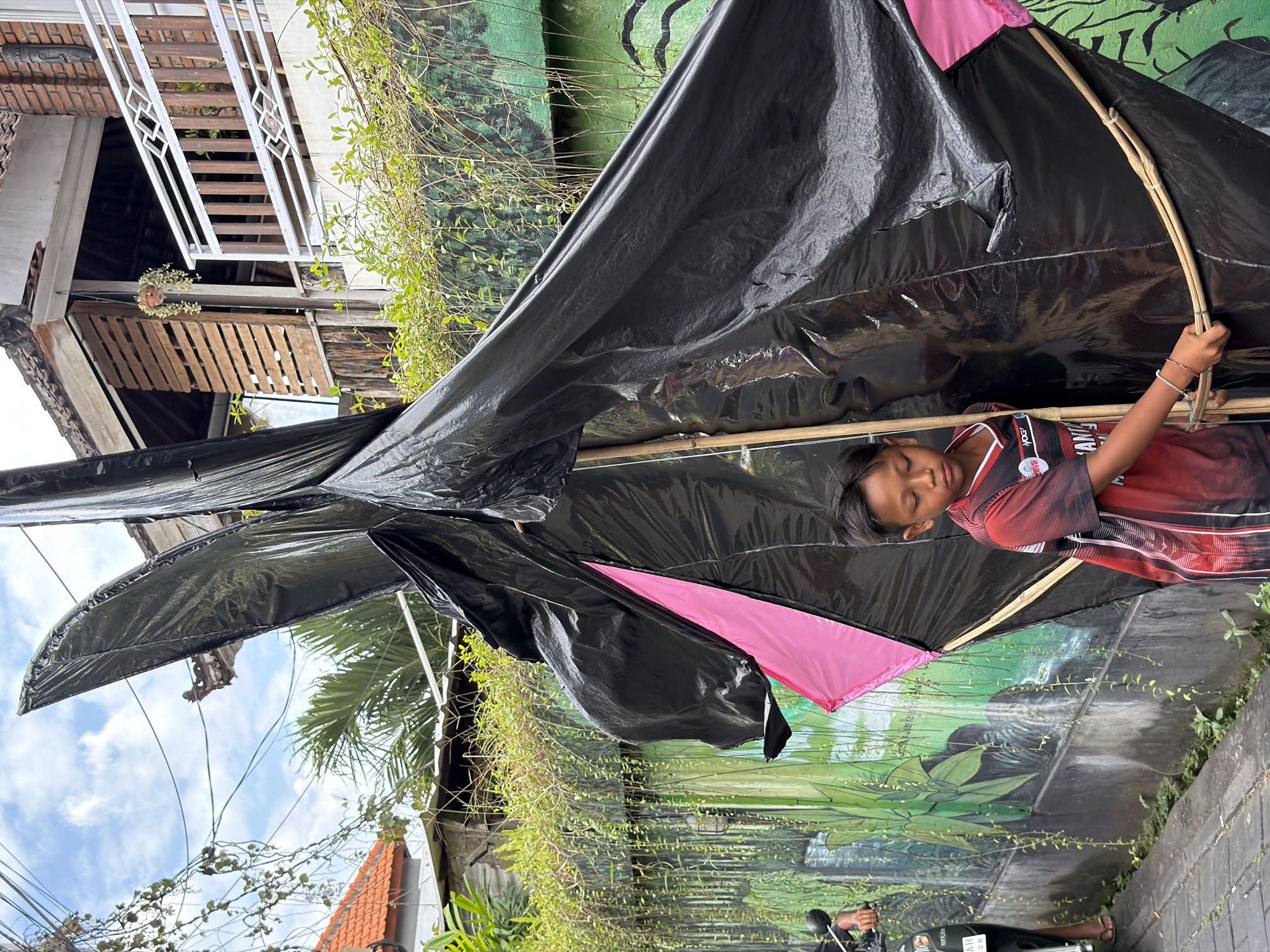 A young boy stands in front of a colorful mural and a large, black kite in an outdoor setting, possibly a street or pathway. The boy, wearing a red and black t-shirt, is looking down at the kite, which is made of black plastic and has pink accents. He is holding onto one of the kite's bamboo frame pieces. To the left of the boy, a portion of a motorcycle and the legs of a person wearing flip-flops are visible. The background includes lush green vines hanging down a wall, a glimpse of a building with a tiled roof, and a sky with scattered clouds. The scene appears to be in a tropical or subtropical location.