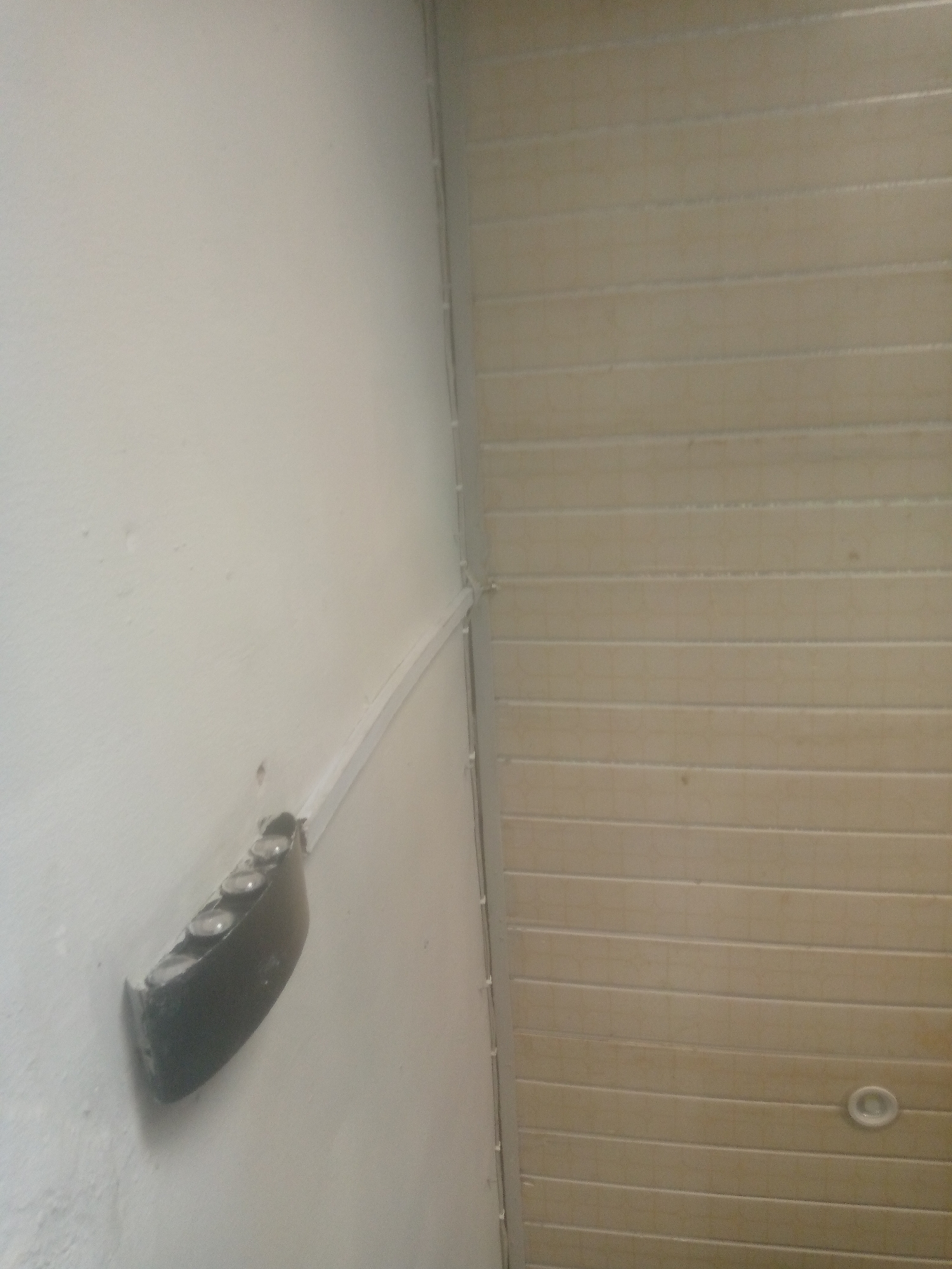 The image shows a close-up view of a white wall with a black electrical extension cord with multiple sockets mounted on it. The extension cord is positioned at an angle and appears to be slightly detached from the wall in some areas. To the right of the wall, a light-colored, horizontally slatted ceiling is visible. There is also a small circular light fixture embedded in the ceiling. The scene is indoors and appears to be a residential or commercial space in Jalingo, Nigeria. There are no people or animals visible in the image. The lighting is neutral, and there are no obvious signs of damage or disrepair other than the slight detachment of the extension cord.