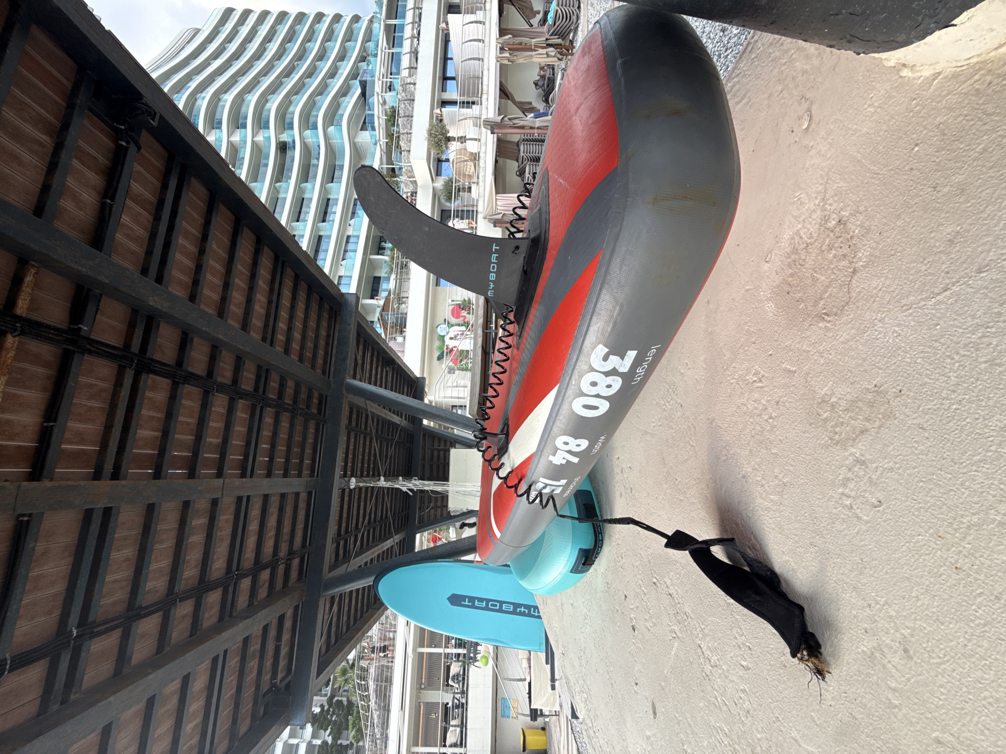 The image shows two paddleboards resting on a sandy surface, likely a beach. The paddleboards are oriented vertically in the frame. The larger paddleboard is red and gray with black accents, and the text "MYBOAT 380 84 length" is visible on its side. It has a black fin attached to its underside. A coiled black leash extends from the fin to a black strap on the ground. A smaller, light blue paddleboard is partially visible beneath the larger one, with the text "MYBOAT" also visible on its side.
In the background, a modern, wavy-facade building with many balconies rises high into the sky. The sky appears overcast, suggesting a cloudy day. The setting appears to be a resort area, with what looks like lounge chairs and umbrellas visible behind the paddleboards and near the building. The overall impression is a relaxed, possibly vacation-oriented scene. No people are clearly visible in the image. The lighting suggests it is daytime. The location cues point to a seaside or waterfront area, with the building and lounge chairs typical of a resort.
