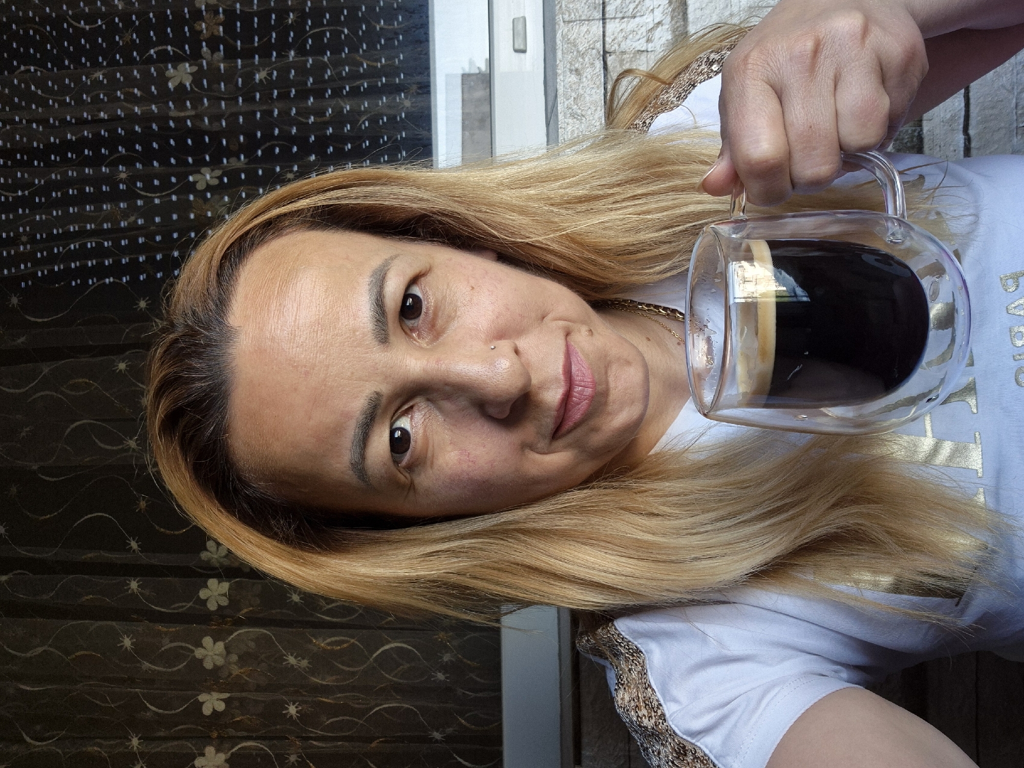 A medium shot shows a woman with blonde hair holding a clear glass mug filled with black coffee. The woman is looking directly at the camera and smiling. She is wearing a white t-shirt with gold lettering on it. The background is a dark curtain with a floral pattern. The scene appears to be indoors, near a window.