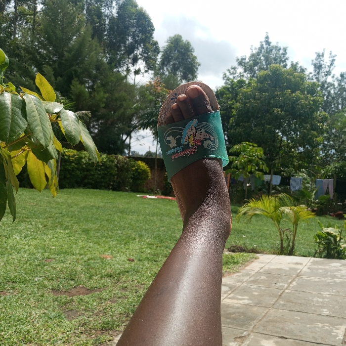 The image depicts an outdoor scene in Mahiakalo ward, Kenya. A dark-skinned human left leg and foot are prominently featured in the foreground, angled upward. The foot wears a light teal slide sandal with a graphic of a surfing character and accompanying text. The immediate ground consists of a light-colored tiled patio adjacent to a manicured green lawn. Various green plants, bushes, and trees populate the midground and background. Distant laundry hangs on a visible clothesline. Tall trees and a partly cloudy sky are present in the background. The scene is illuminated by natural daylight, with highlights on the skin.