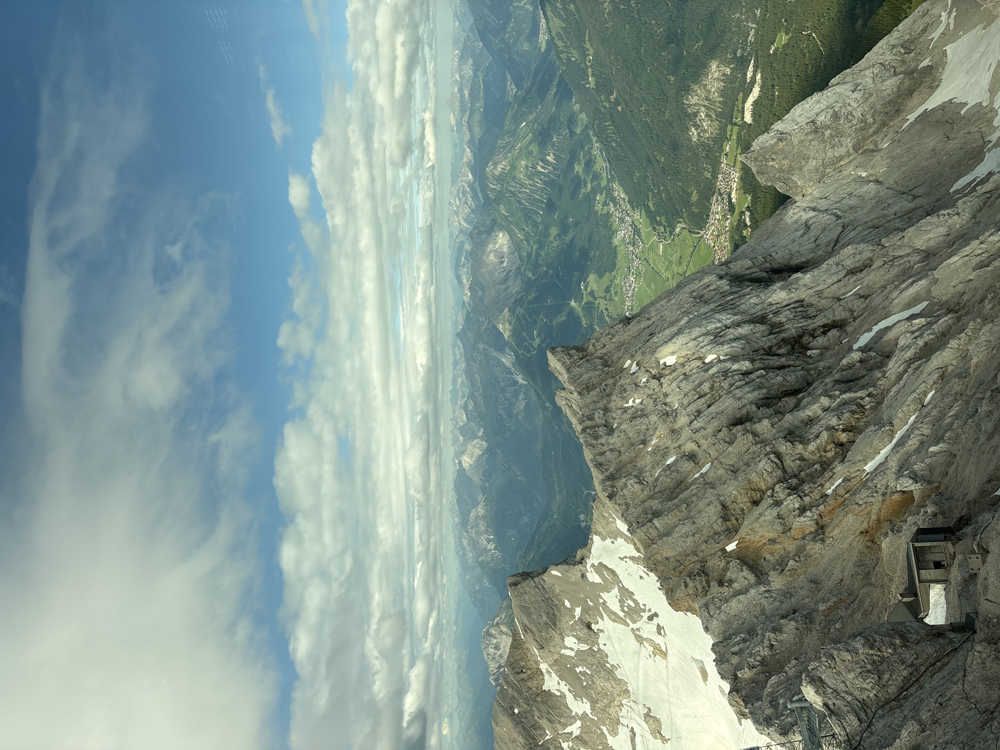 This image is a wide, high-angle, vertical shot of a rugged mountain landscape. In the foreground, a steep, rocky mountain face dominates the left side of the frame, with patches of snow clinging to its crevices. A small, dark, hut-like structure is visible nestled into the rock face near the bottom left. The mountain slopes downwards into a verdant valley dotted with buildings and trees, indicative of a town or village. This valley is framed by layers of receding mountain ranges that stretch into the distance. The sky above is a vibrant blue, filled with scattered, fluffy white clouds. The scene depicts a dramatic natural environment with signs of human habitation in the valley below. The viewpoint suggests the photo was taken from a very high elevation, possibly a mountaintop or a cable car, looking down and across the valley. The presence of a town in the valley and the distinct mountain ranges are consistent with the location of Ehrwald, Austria.